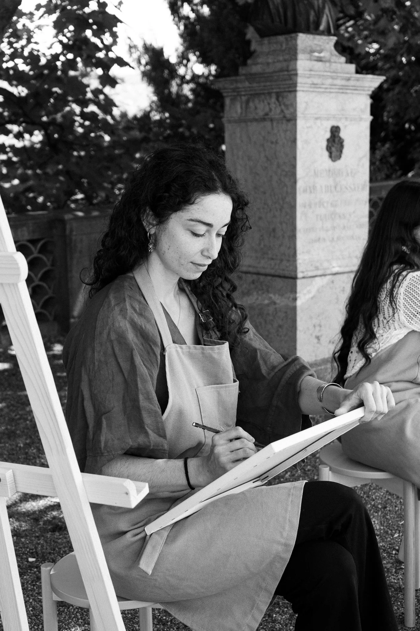 A woman with curly black hair sitting on a bench outdoors under a tree, painting on a canvas, surrounded by flowers and greenery.