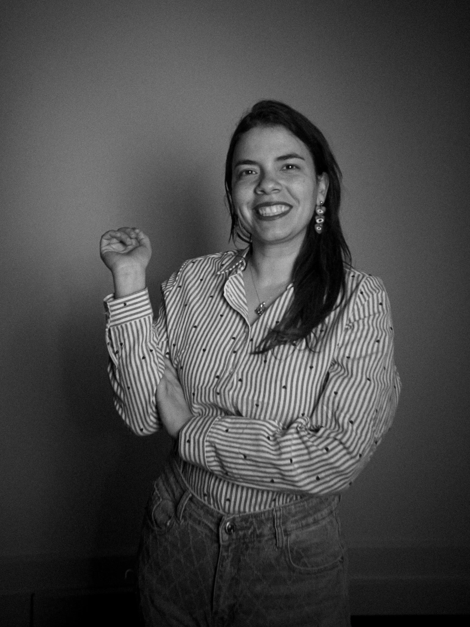 A woman with long dark hair smiling, wearing a striped shirt with small polka dots and red earrings, standing against a gradient background.