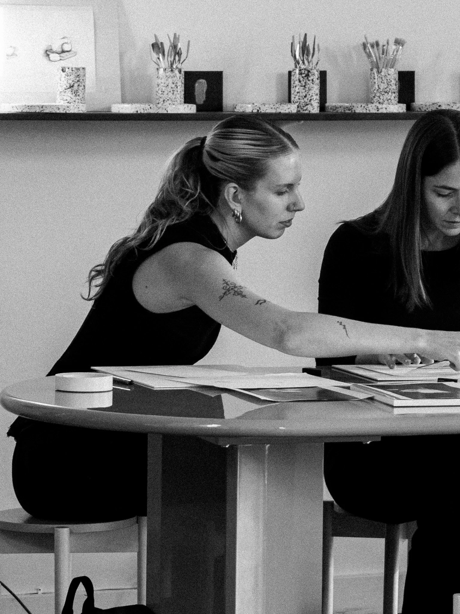 A young woman with blonde hair, tattoos on her arms, and a black sleeveless top sitting at a table in a well-lit room, drawing on paper with a pen. The room has large windows and shelves with ceramic items in the background.