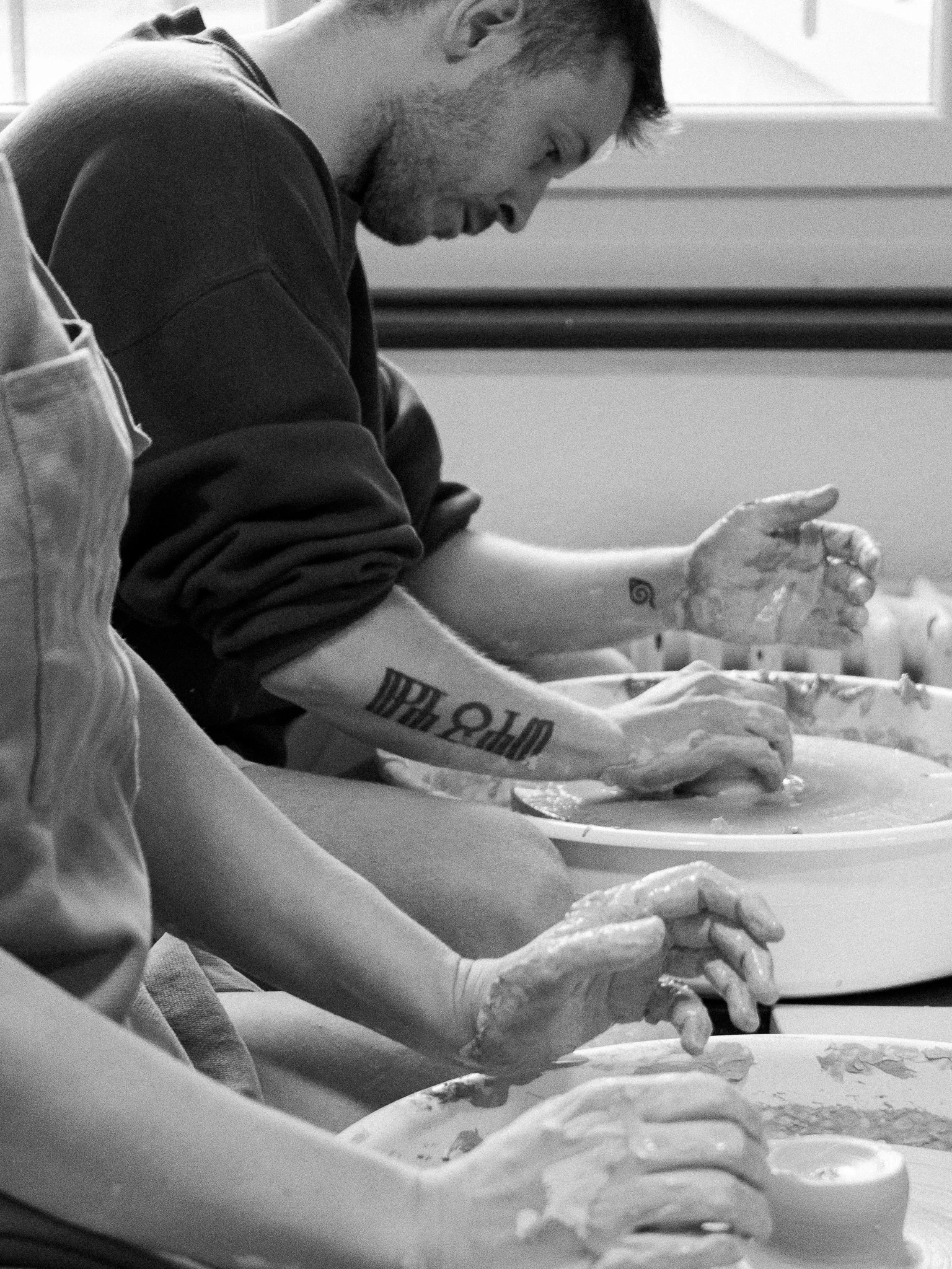 A man with brown hair, beard, and a tattoo on his neck smiling as he molds clay on a pottery wheel. He is wearing a gray apron with orange accents, working in a pottery studio with shelves of pottery supplies in the background.