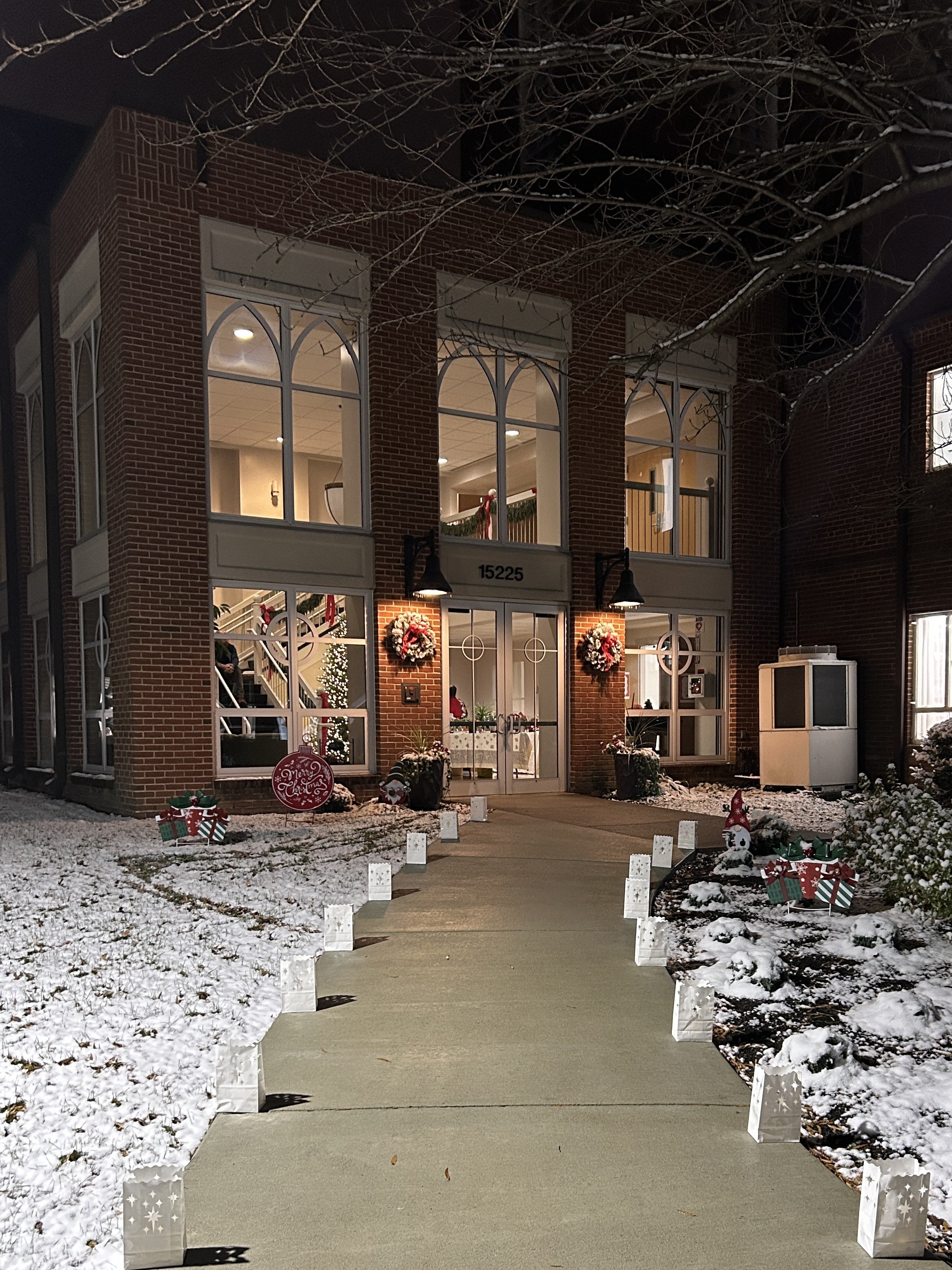 A brick building decorated for Christmas with wreaths and lights, a table with a holiday display inside, and a snow-covered walkway lined with paper lanterns, during nighttime.