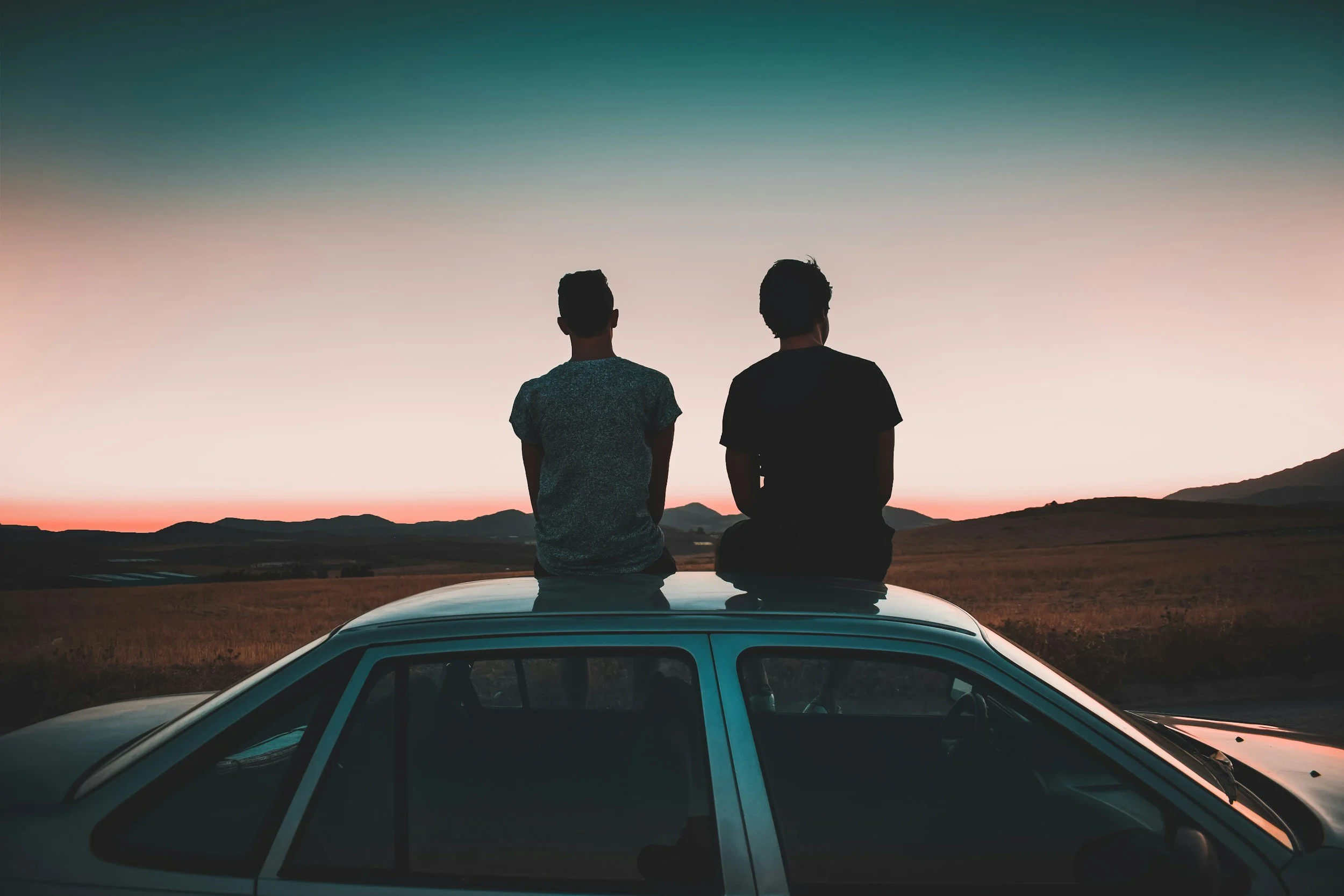 Two people sit on the roof of a car, watching a sunset over a rural landscape with rolling hills and a few scattered trees.