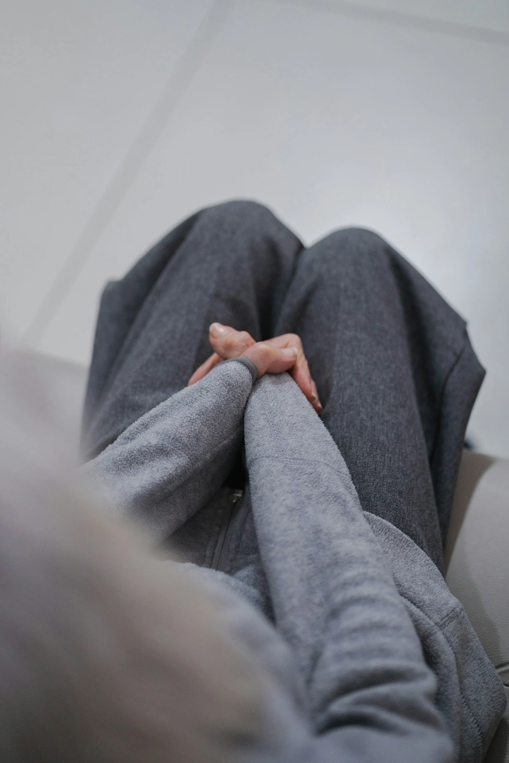 Photo of a person wearing gray pants and a gray hoodie, sitting on a white chair with their hands resting on their lap, holding their fingers intertwined.
