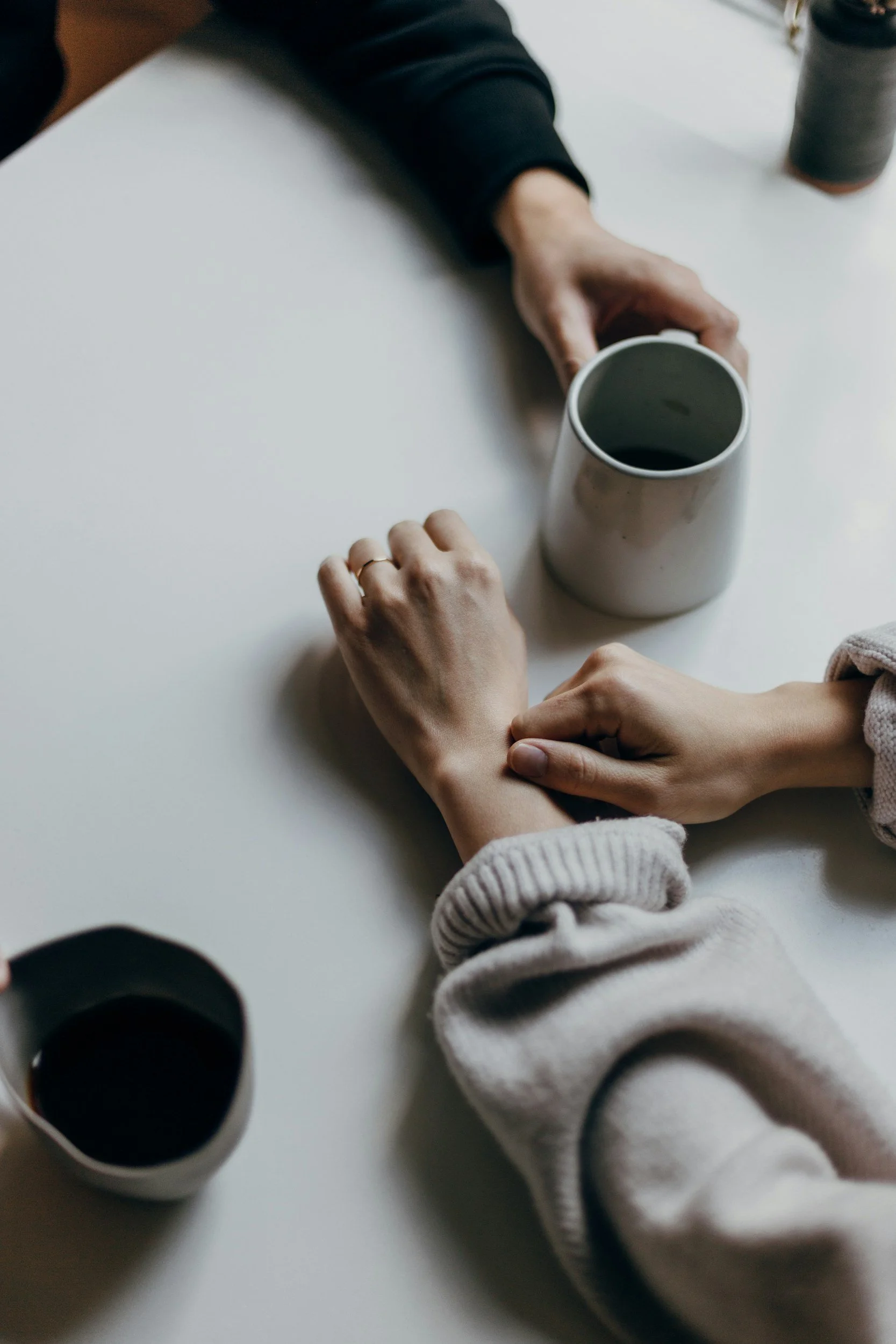 People at a white table with coffee mugs, one person wearing a grey sweater, another in black, holding a mug. Coffee mugs on the table.