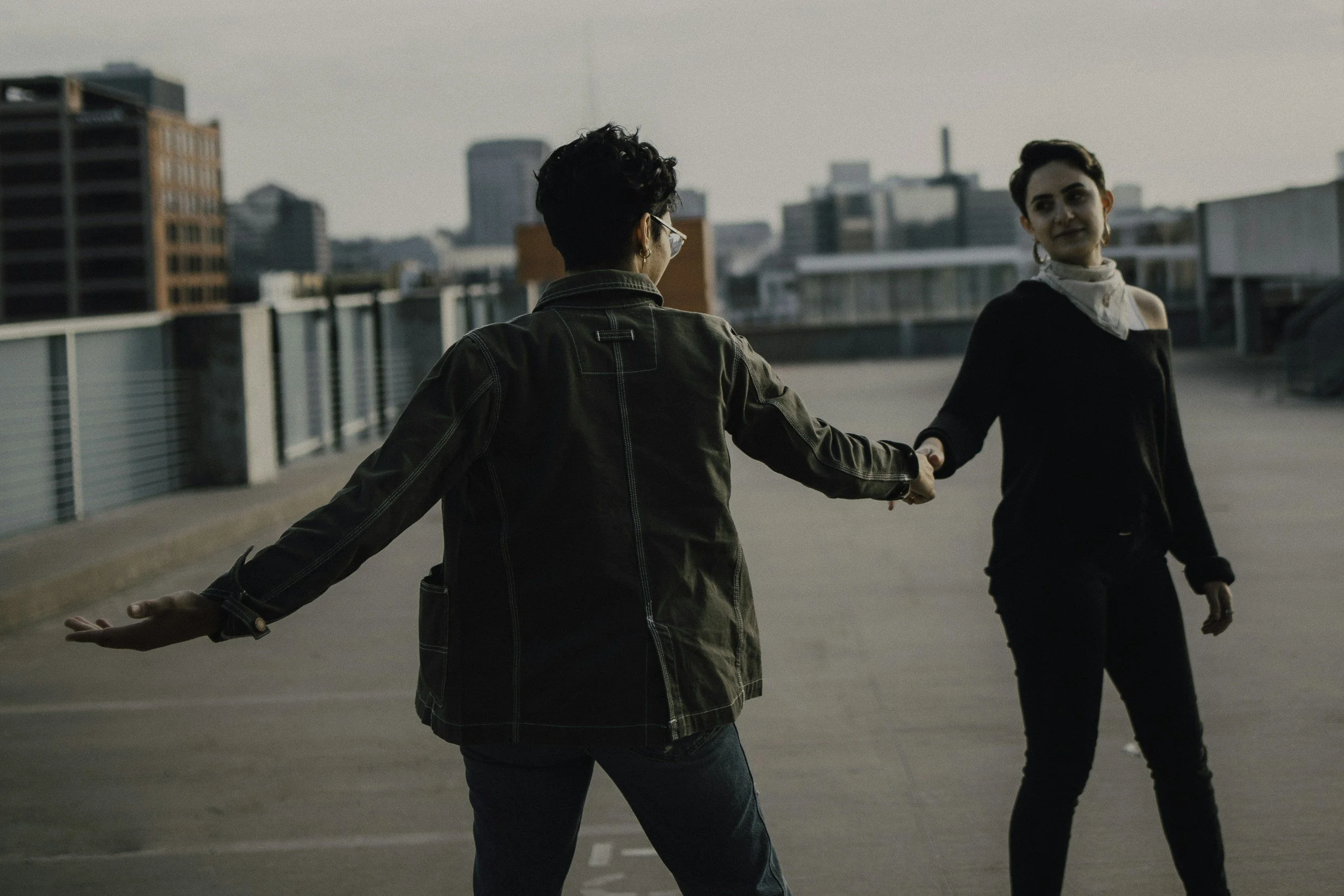 Two women are dancing or swinging on a rooftop in an urban setting, with city buildings in the background during late afternoon or early evening.