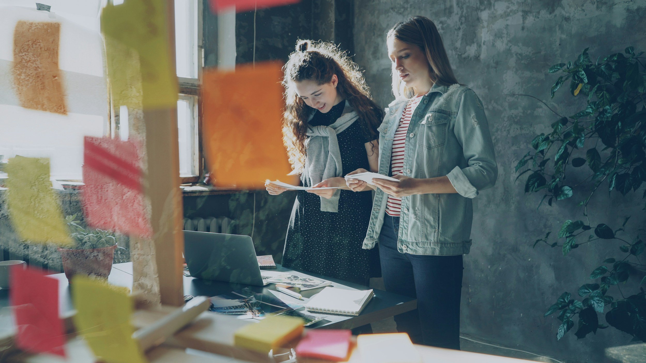 Two young women stand at a table inside an industrial-style room with concrete walls and large windows, looking at papers and smiling. Various office supplies and a laptop are on the table.