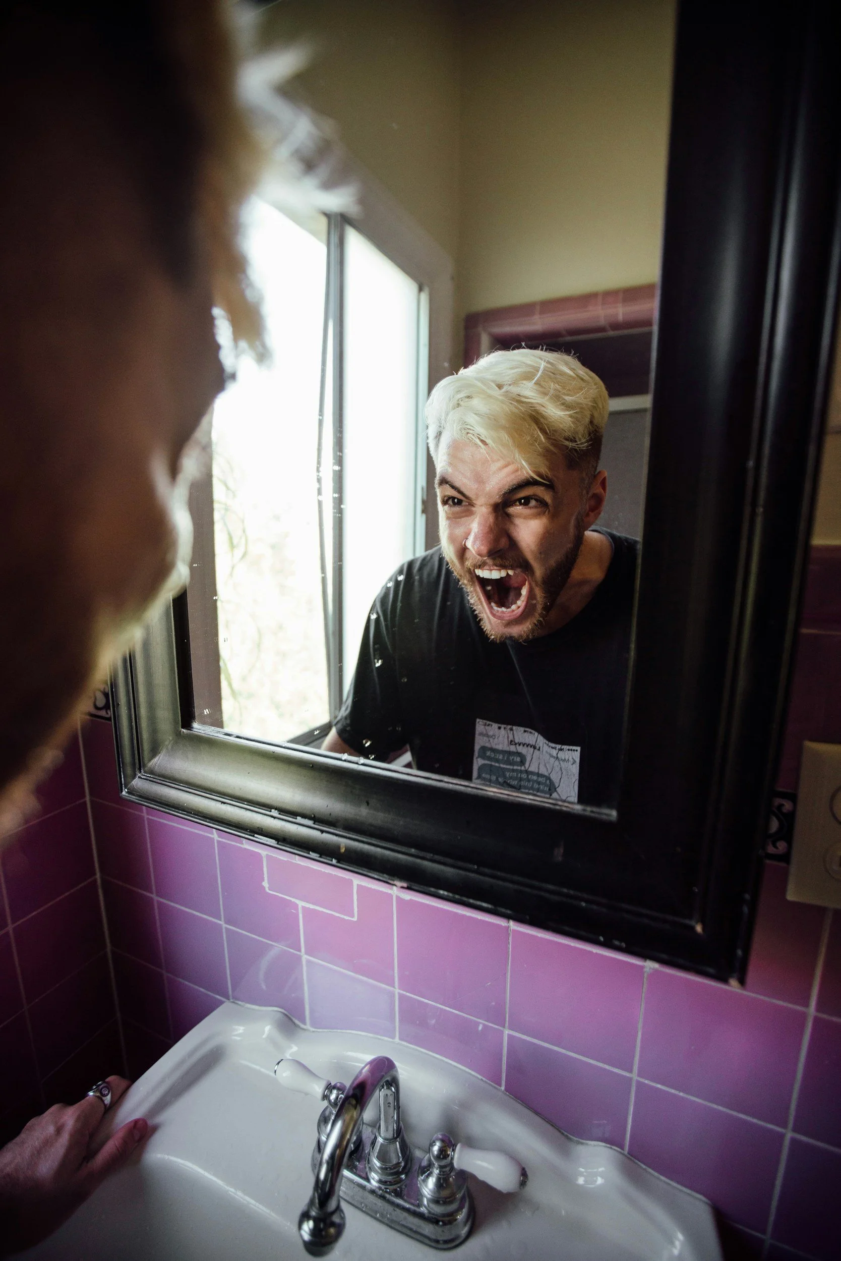 A person with blonde hair and a beard looking angry or shouting, seen through a bathroom mirror. The bathroom has pink tiles, a white sink, and a silver faucet. The person's reflection is partially obscured by the mirror frame.