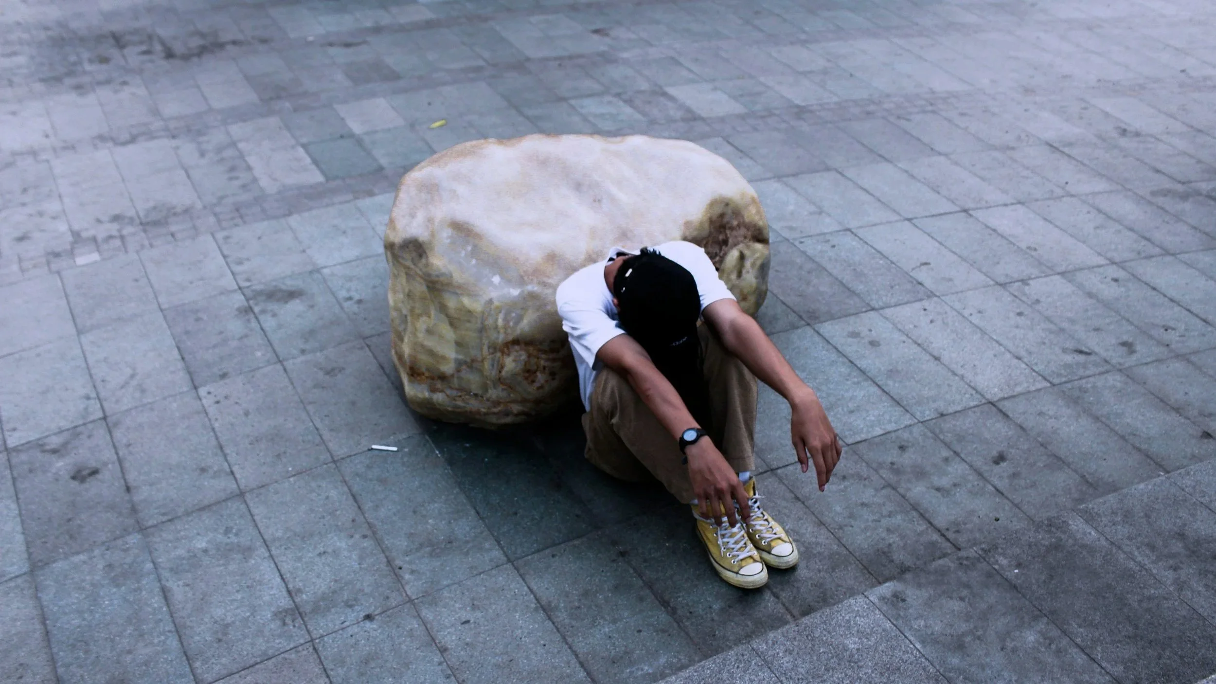 A person sitting on the ground with a large rock behind their back, wearing a black cap, white T-shirt, brown pants, and yellow sneakers, with their head lowered and arms resting on their knees in a public area with tiled pavement.
