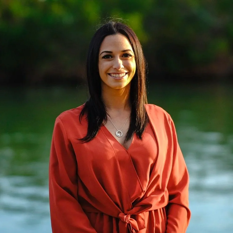 A woman with long dark hair smiling outdoors, wearing an orange wrap dress and a spiral necklace, with a green and water background.