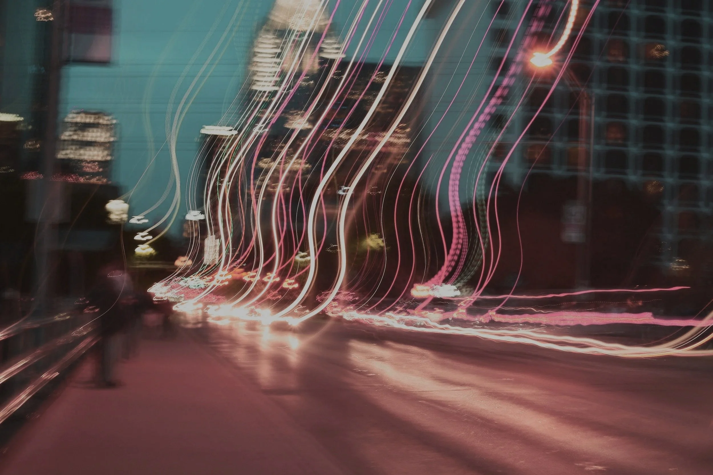 Nighttime city street with light trails from moving vehicles and buildings in the background.