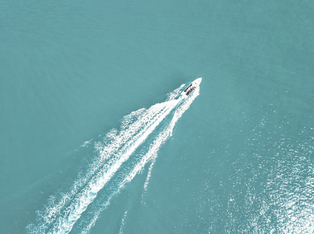 A white motorboat moving at high speed on a turquoise body of water, creating a spray of water behind it and leaving a foamy wake.
