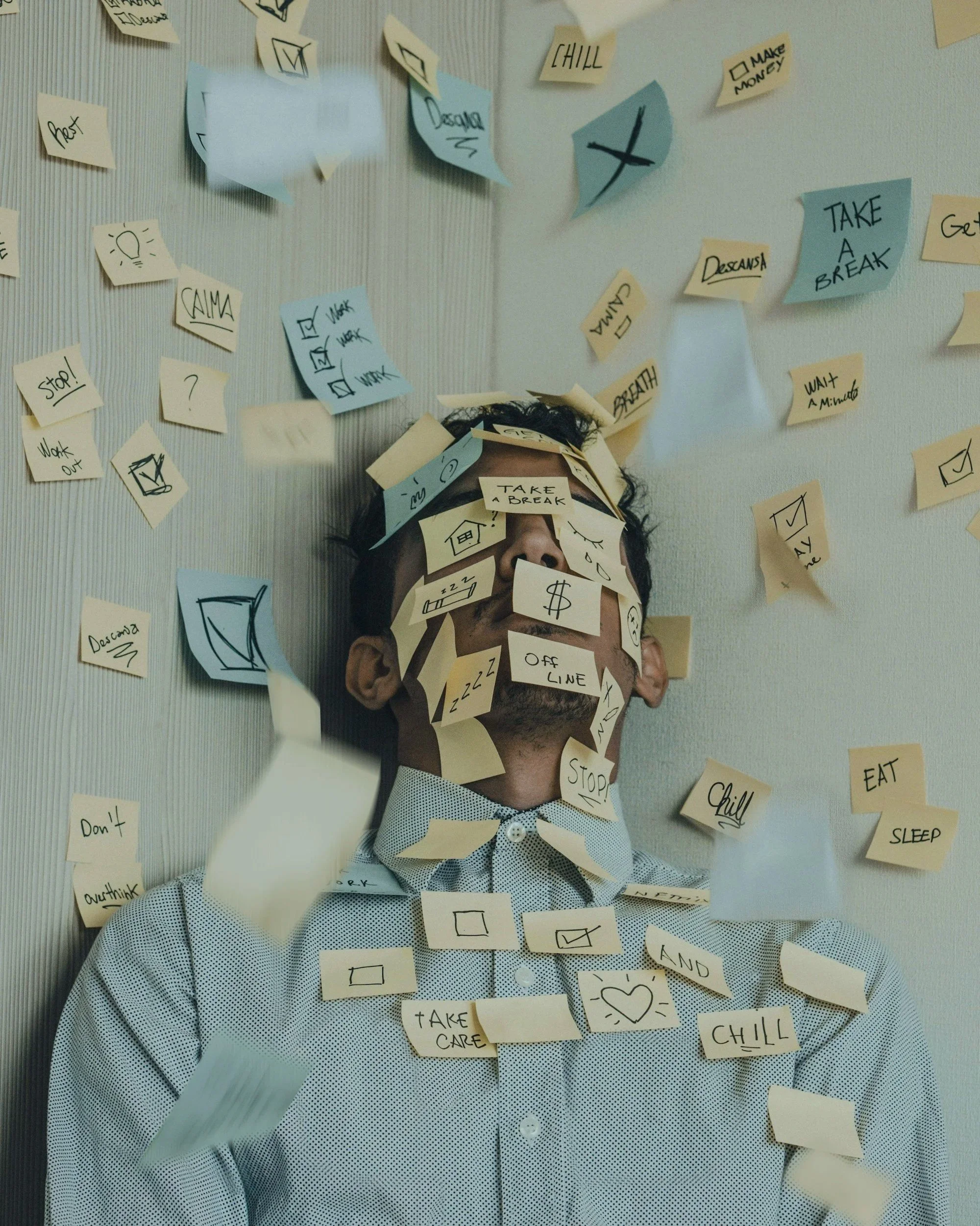 Person with short dark hair and wearing a light-colored, patterned button-up shirt, surrounded by yellow and blue sticky notes with handwritten notes and symbols, some attached to their face and others on the wall, creating a chaotic scene.