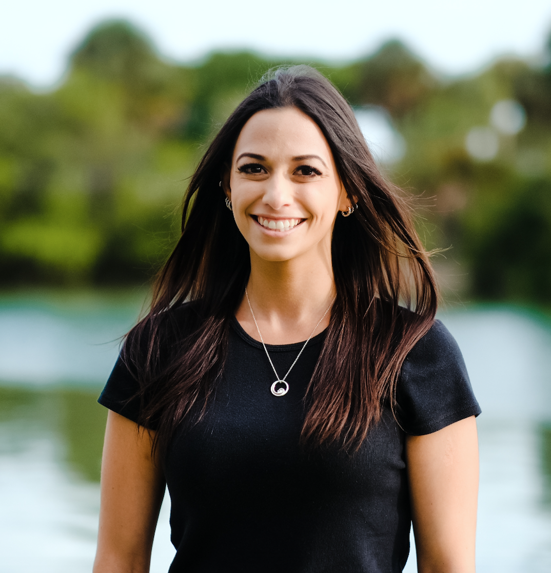 A woman with long dark hair smiling outdoors with a blurred green and blue background.