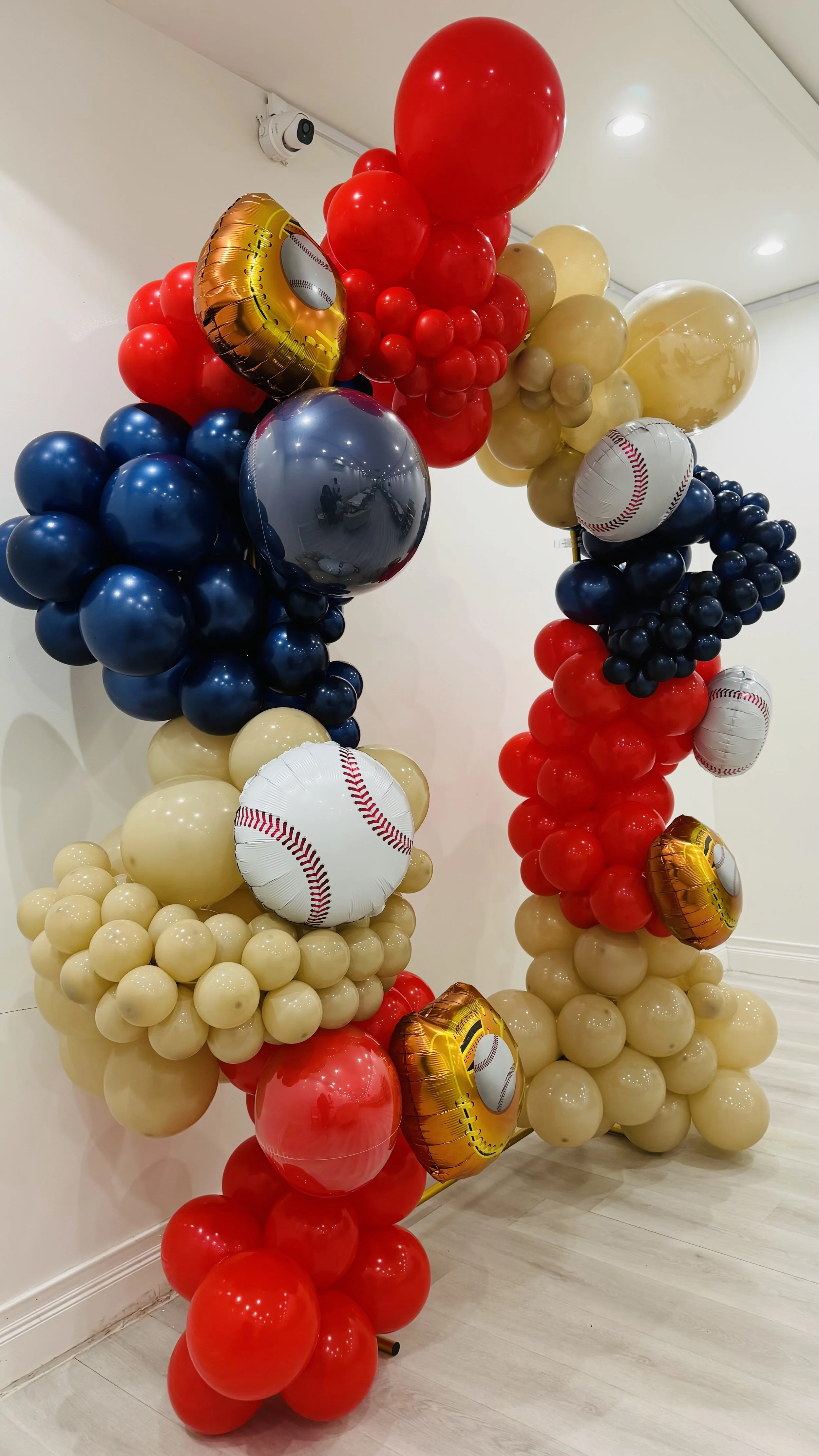 Colorful balloon arrangement with red, beige, navy blue, and metallic gold balloons, including baseball-themed balloons with baseball designs.