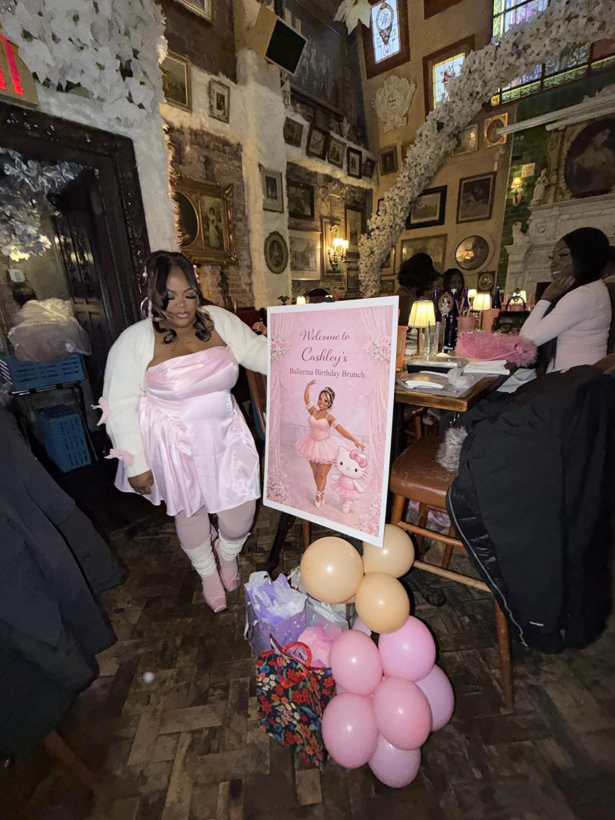 A woman dressed in pink ballerina costume at a birthday celebration, with pink and beige balloons decorating the area, and a sign that says 'Welcome to Cashley's Ballerina Birthday Brunch'.