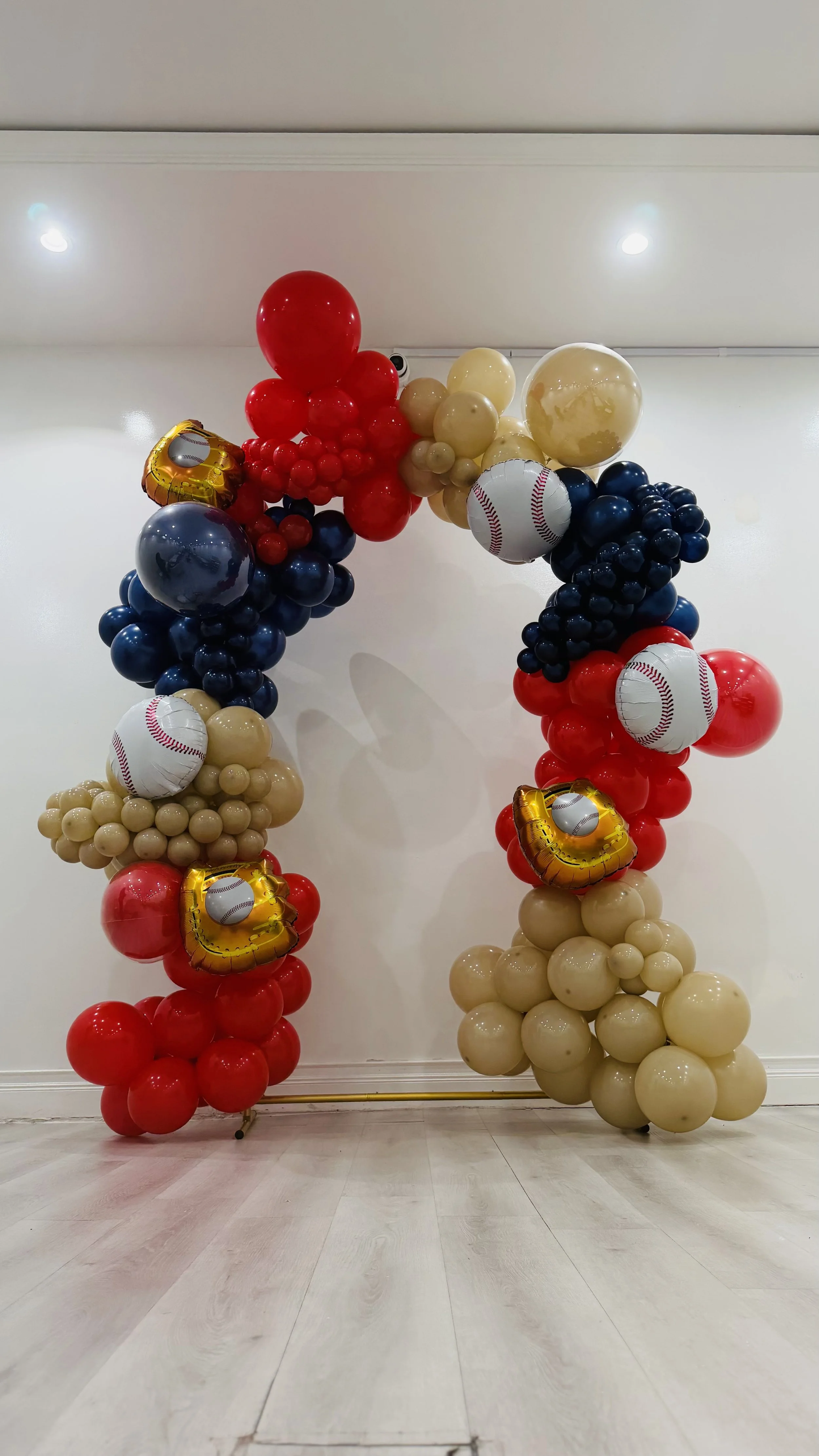 Balloon arch with red, navy, beige, and gold balloons, featuring baseball-themed balloons and gold heart-shaped balloons, set against a white wall and light wooden floor.