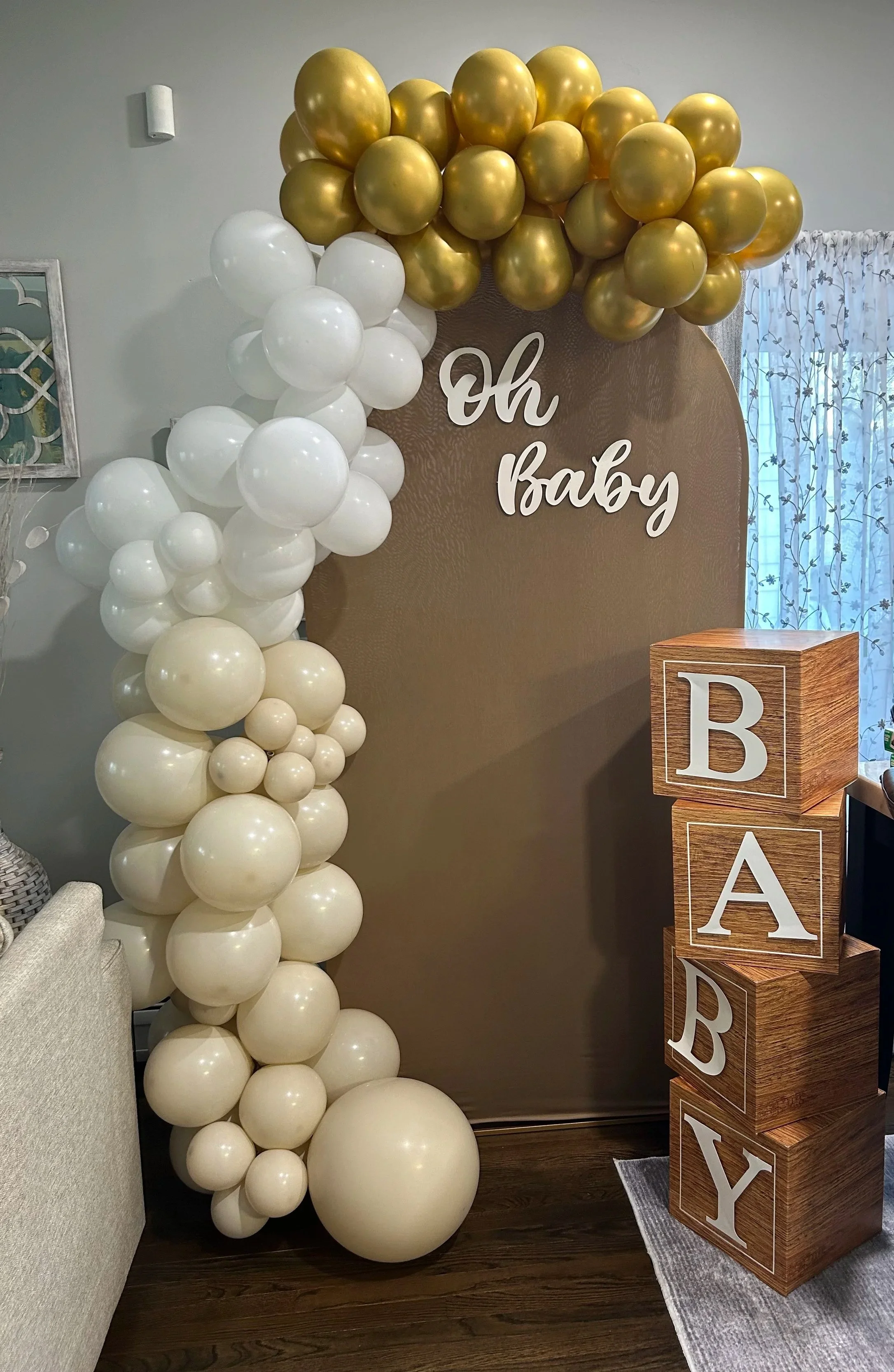 Decorative balloon arch with white and gold balloons and a backdrop with the words "Oh Baby" written on it. Wooden boxes with large letter blocks spelling "BABY" are stacked nearby.