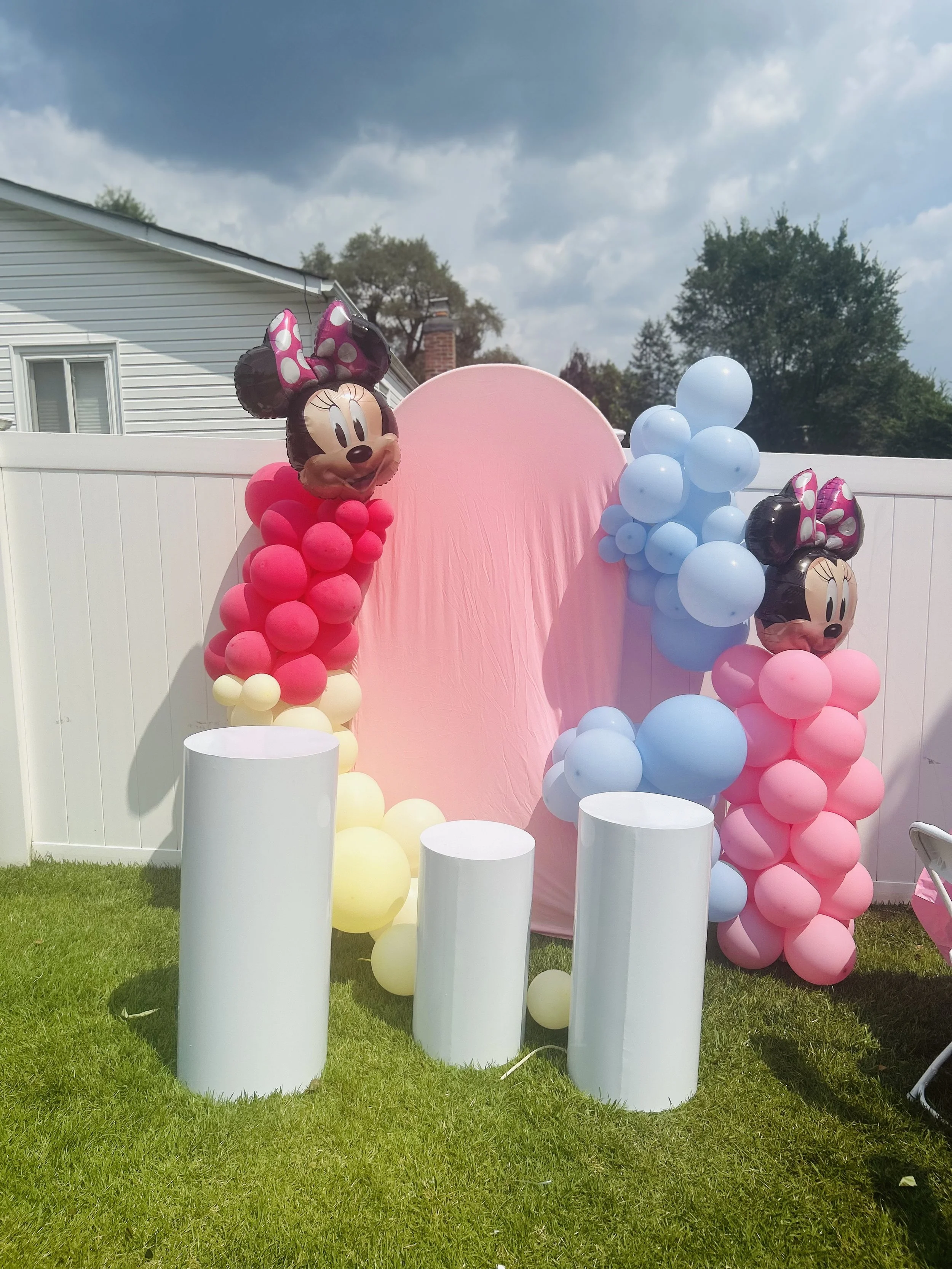 Decorative backdrop for a birthday celebration featuring pink and blue balloons arranged in clusters, with Minnie Mouse shaped balloons on either side, and four white cylindrical pedestals in front on a grassy yard with a white fence.