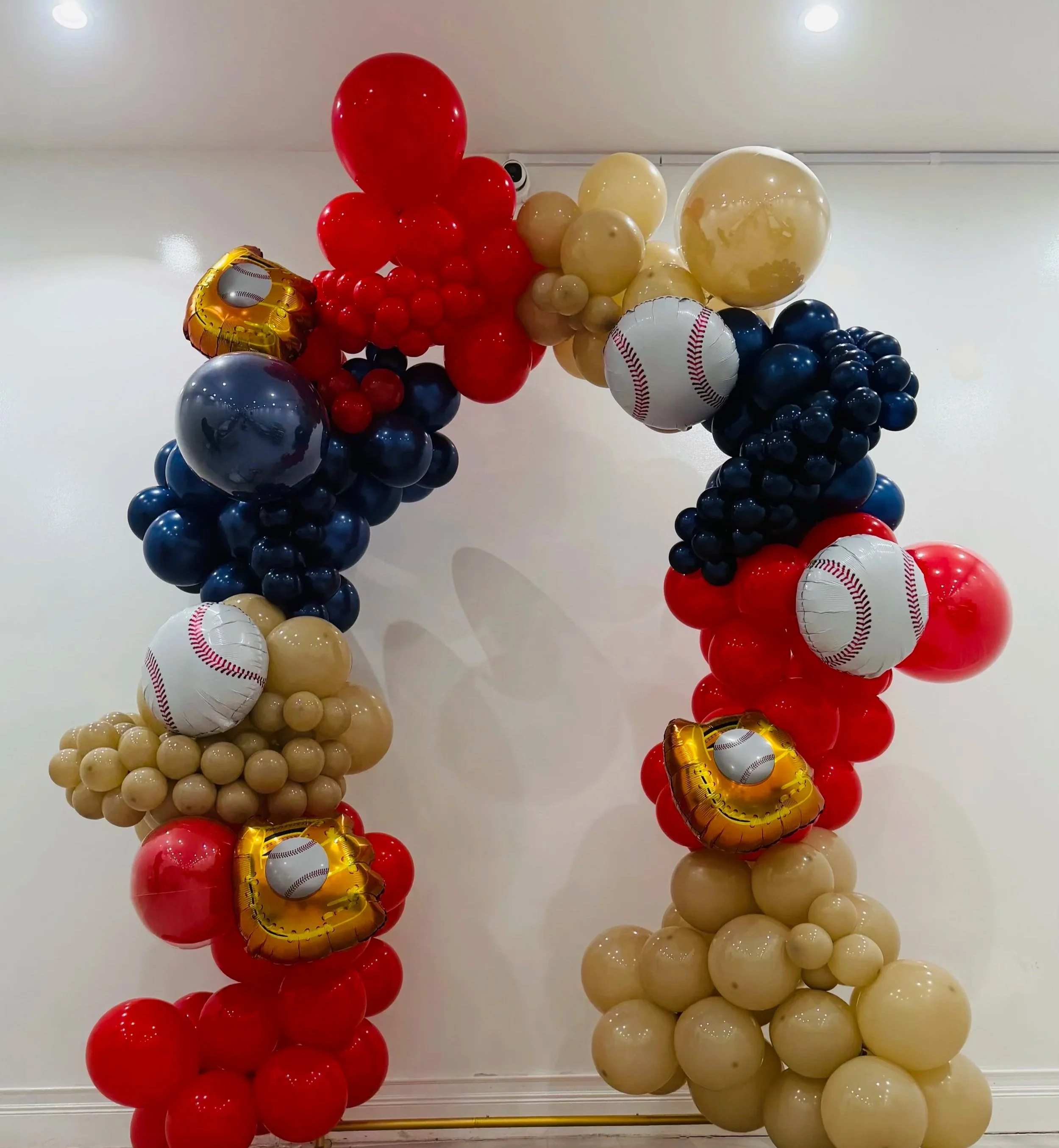 Balloon arch featuring red, cream, navy blue, and gold balloons, some shaped like baseballs, decorated for a sports-themed celebration.