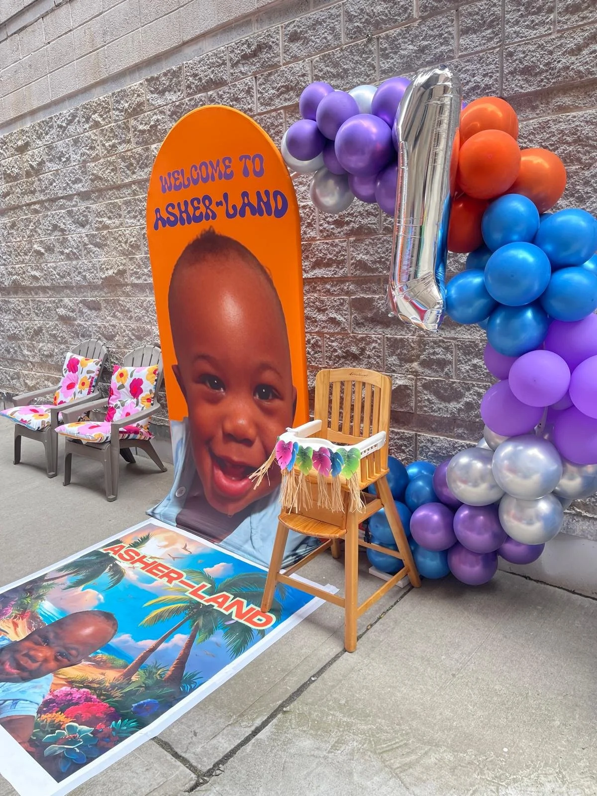 A birthday decoration display featuring a large banner with a child's face and the words "Welcome to Asher-Land," surrounded by colorful balloons in purple, blue, silver, and orange, with a silver number "1" balloon. There are three chairs with flora