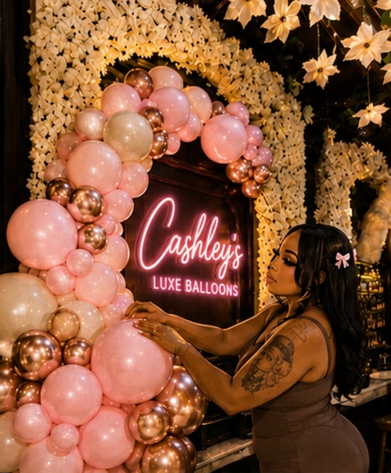 A woman with tattoos arranging pink, gold, and pearl-colored balloons around a neon sign that reads 'Cashley's Luxe Balloons' at a decorated event space with floral arches.