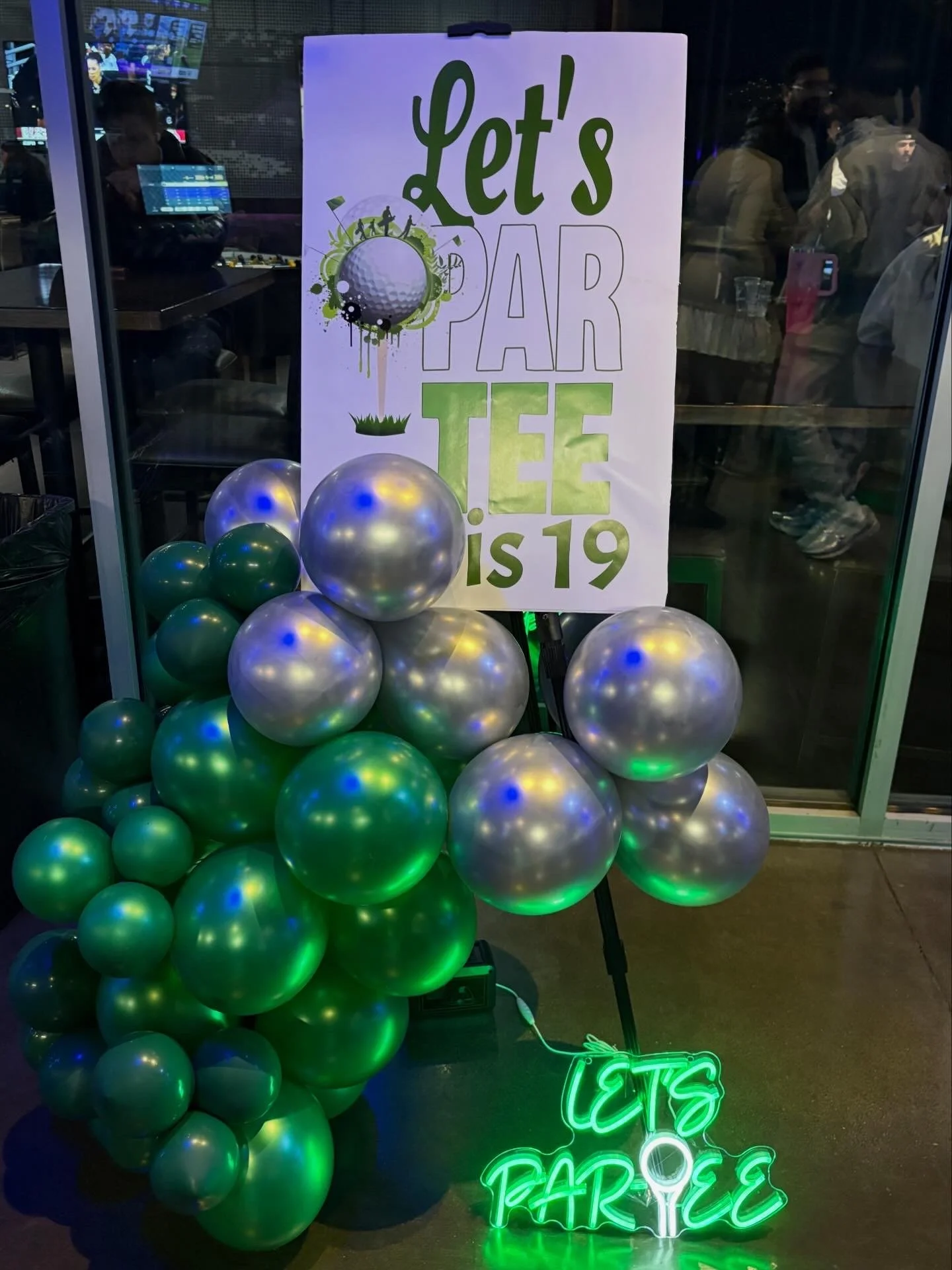 A sign that reads 'Let's Par-Tee! is 19' with a golf ball graphic, surrounded by green and silver balloons, and a neon sign that says 'Eat, Drink, Play' in green near the bottom.