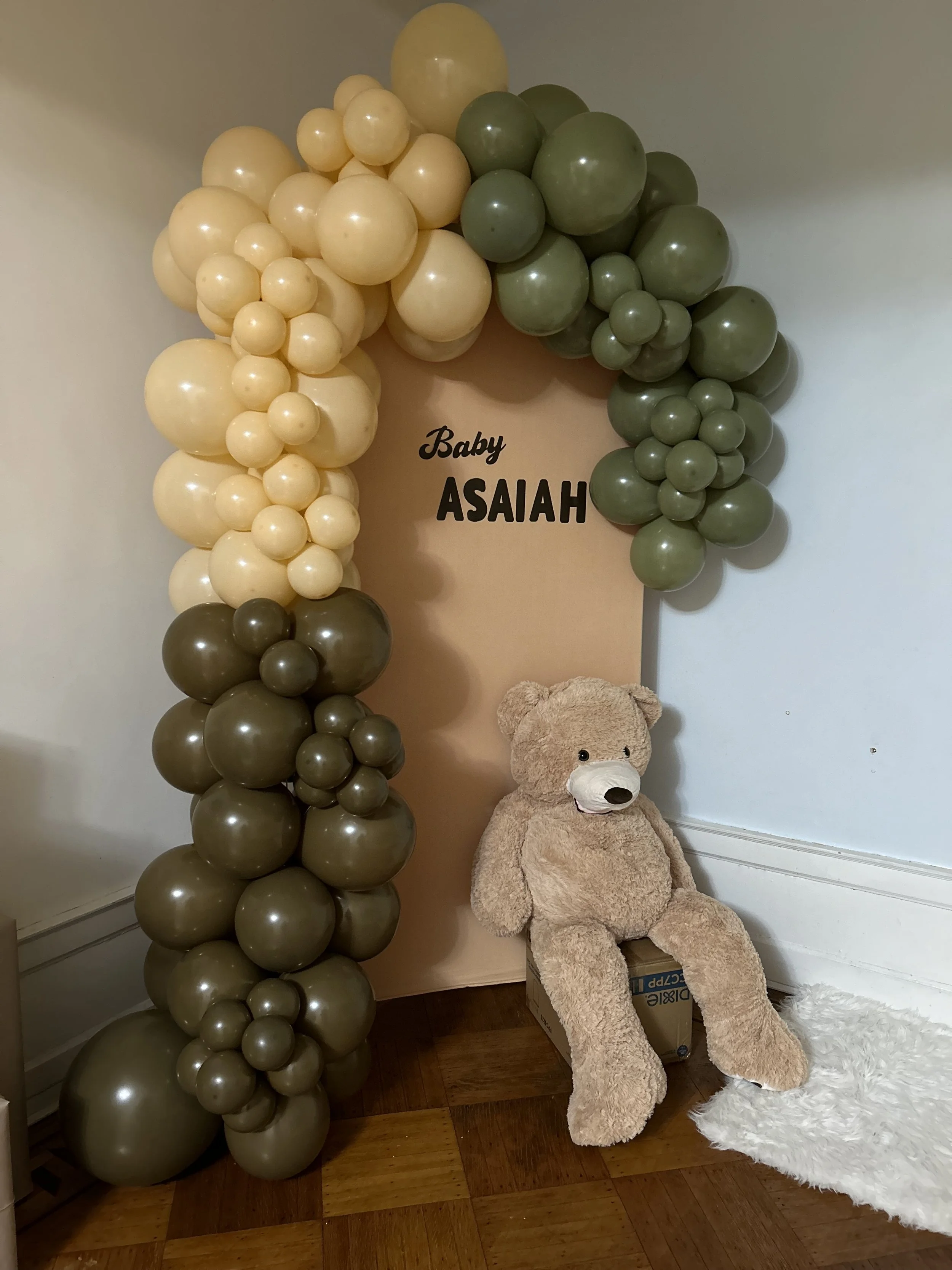 Balloon arrangement with cream and green balloons, a beige teddy bear sitting on a box, a white fluffy rug, and a backdrop with the text 'Baby ASAIAH' in black font.
