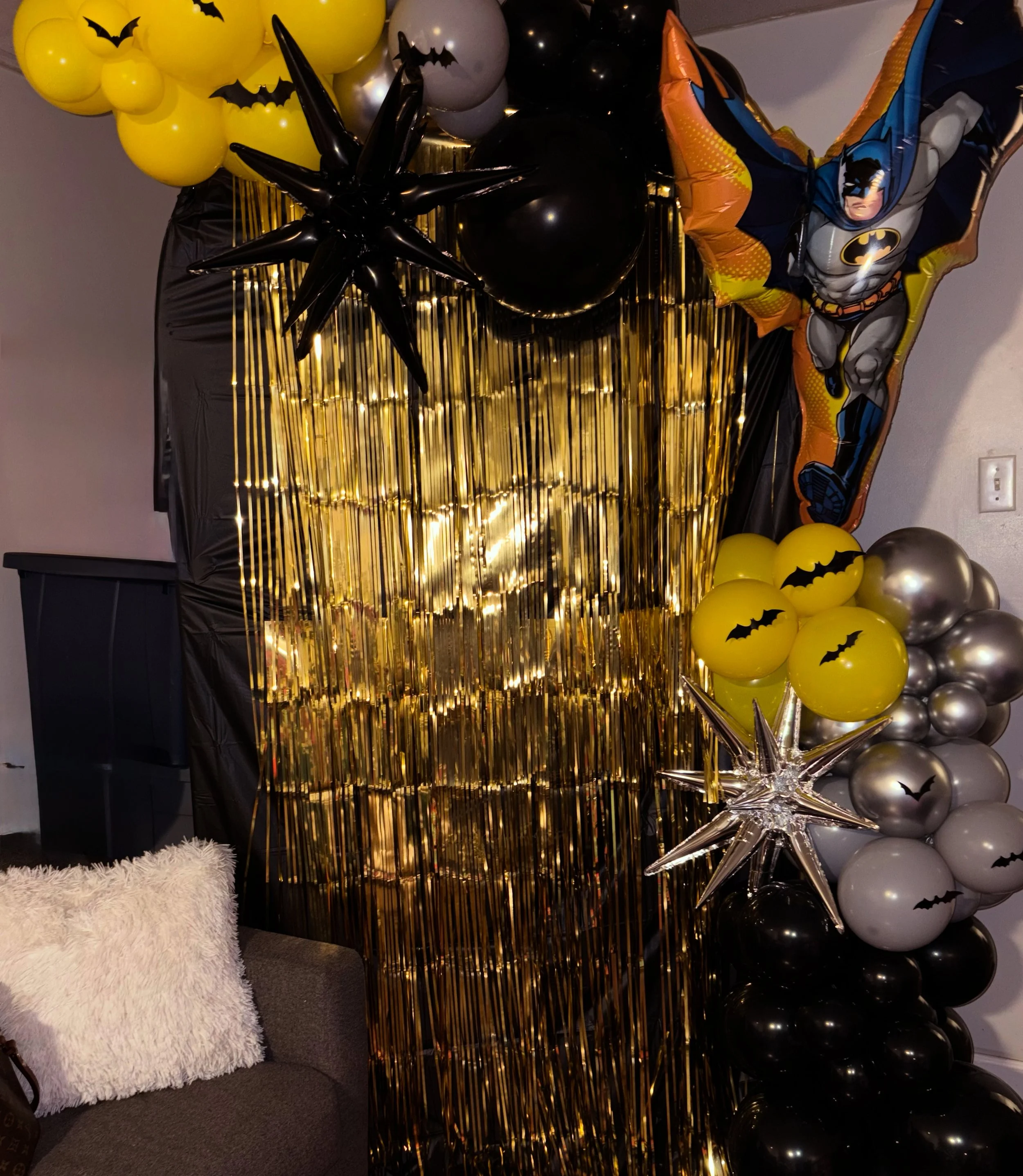 Halloween-themed balloon decoration with yellow and black balloons, some with Batman and bat silhouettes, and a foil balloon of Batman in an action pose. There are black and silver balloons, gold tinsel backdrop, and a large silver star-shaped balloo
