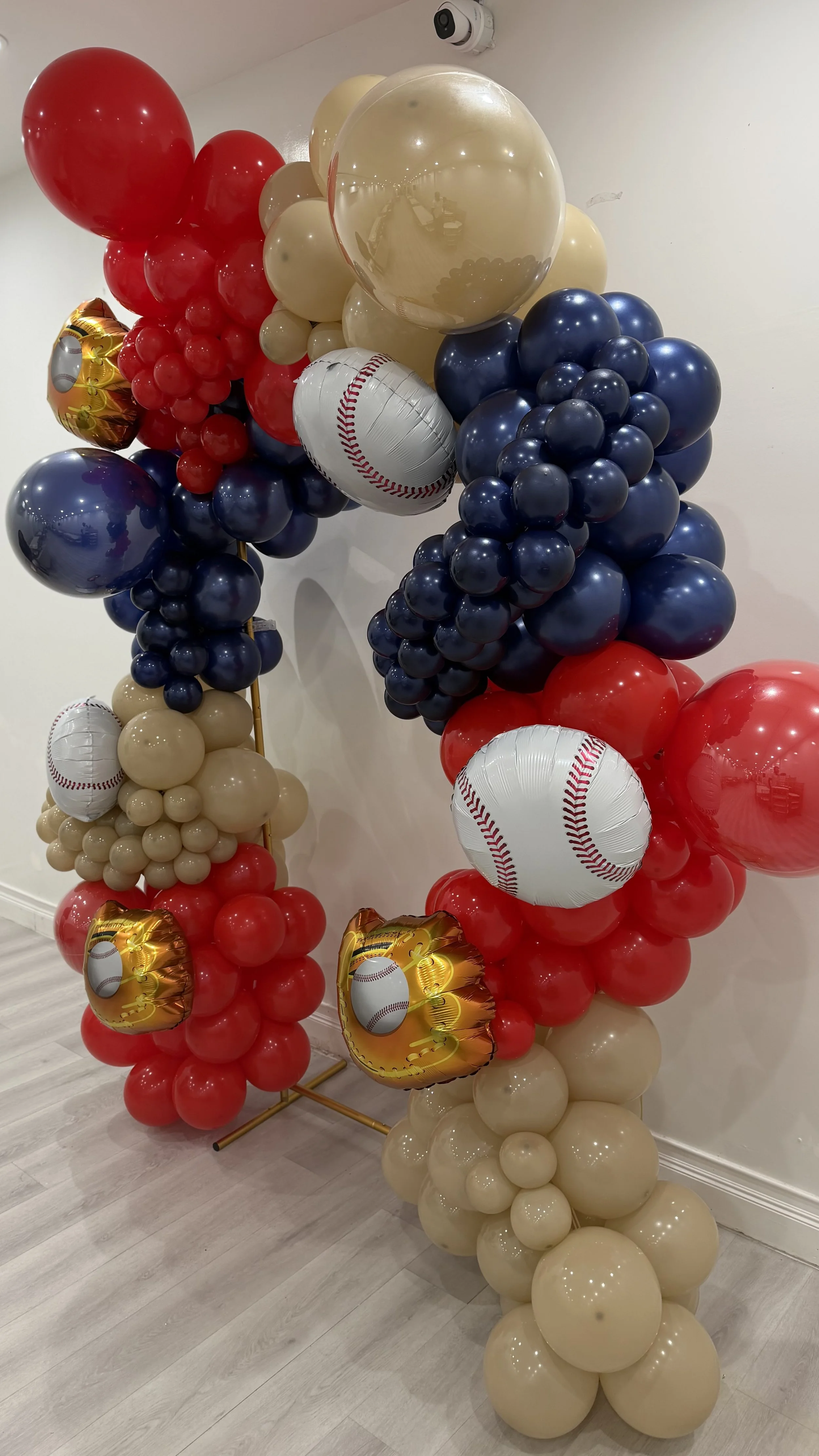 Balloon display with red, beige, blue, and baseball-themed balloons, arranged on a gold stand.