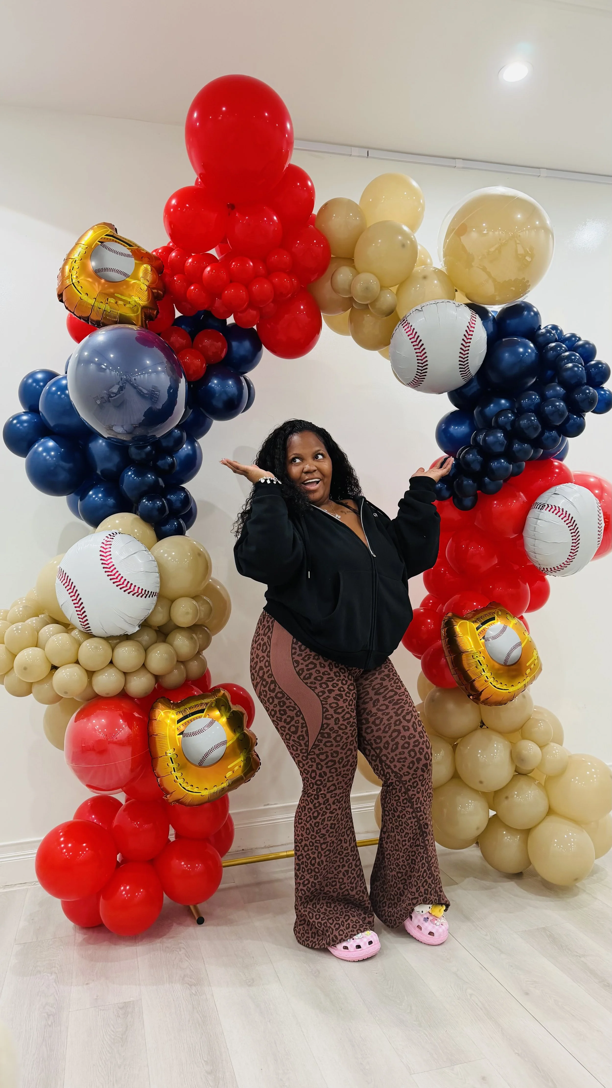 A woman posing under a baseball-themed balloon arch made of red, blue, beige, and gold balloons, with some shaped like baseballs and others as gold and red clusters, indoors with white walls and wooden floor.