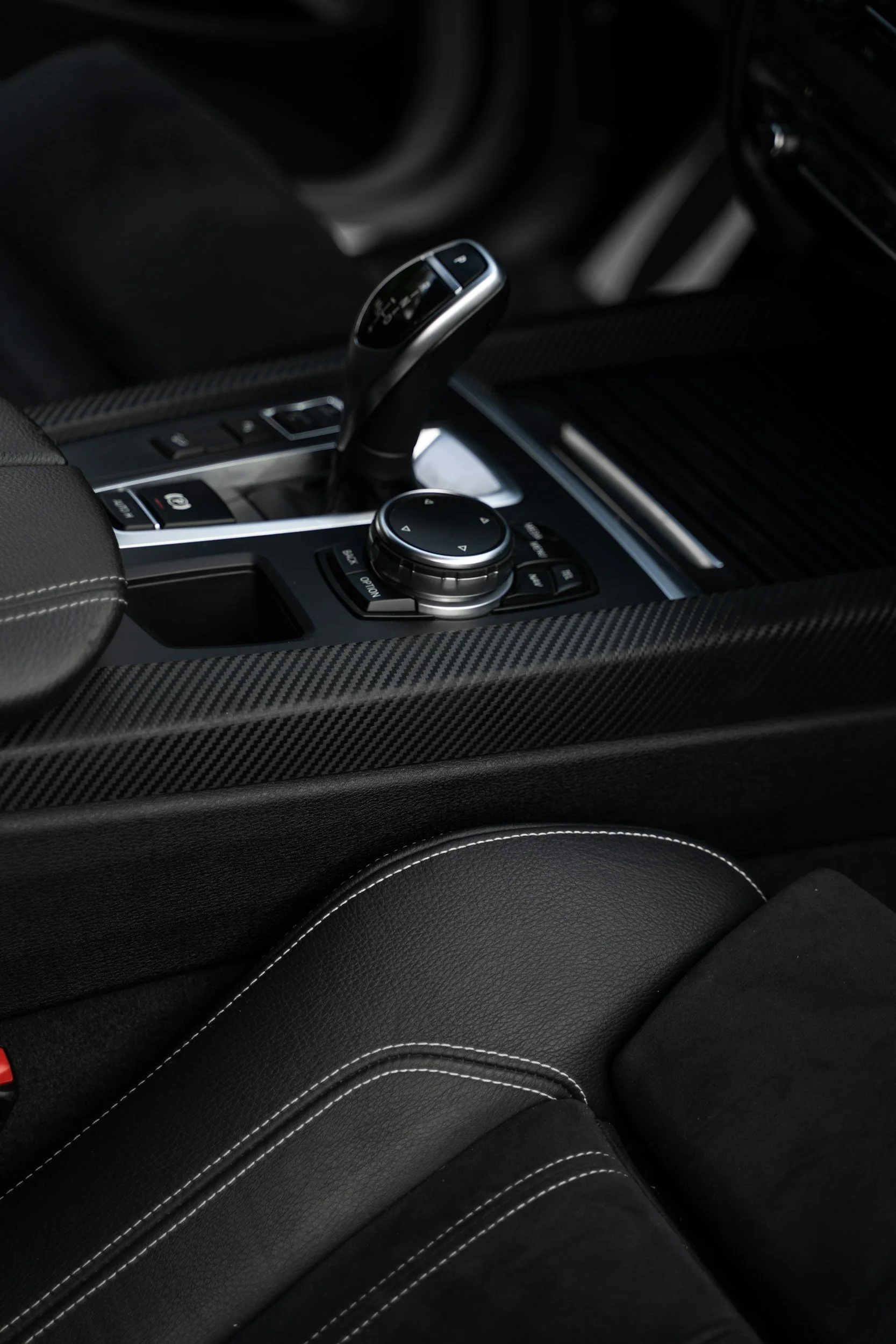 Close-up of a modern car's center console featuring a gear shift, control knob, and buttons, with a carbon fiber trim design.