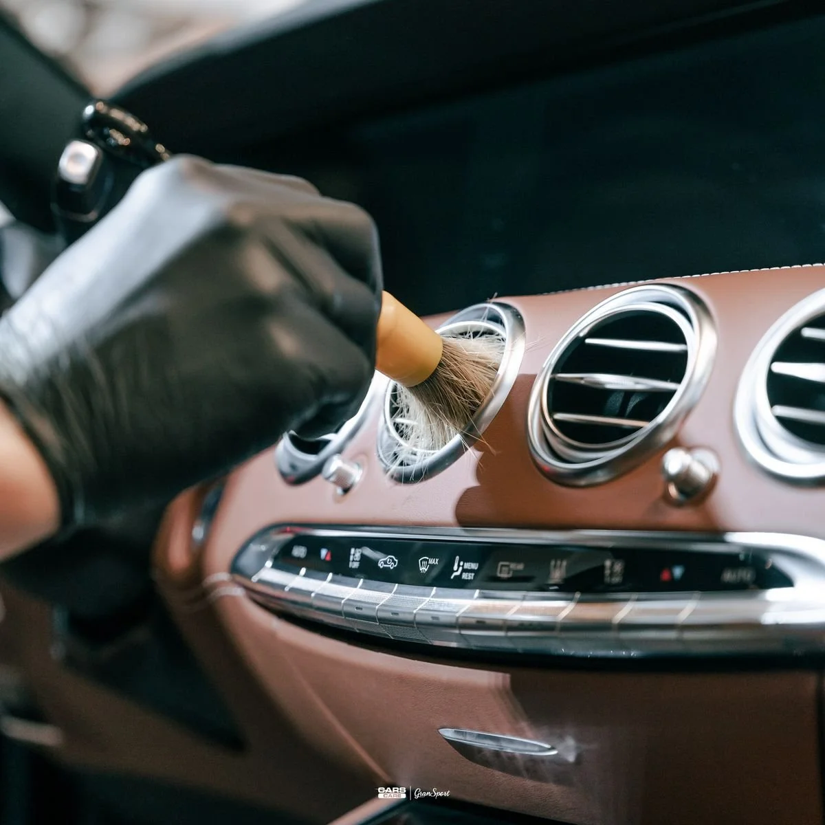Person wearing black gloves cleaning or dusting the air vents and dashboard of a car with a small brush.