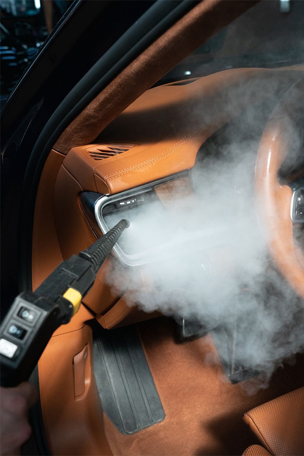 A person using a steam cleaner on the dashboard of a car with brown leather interior, creating visible steam and fog.