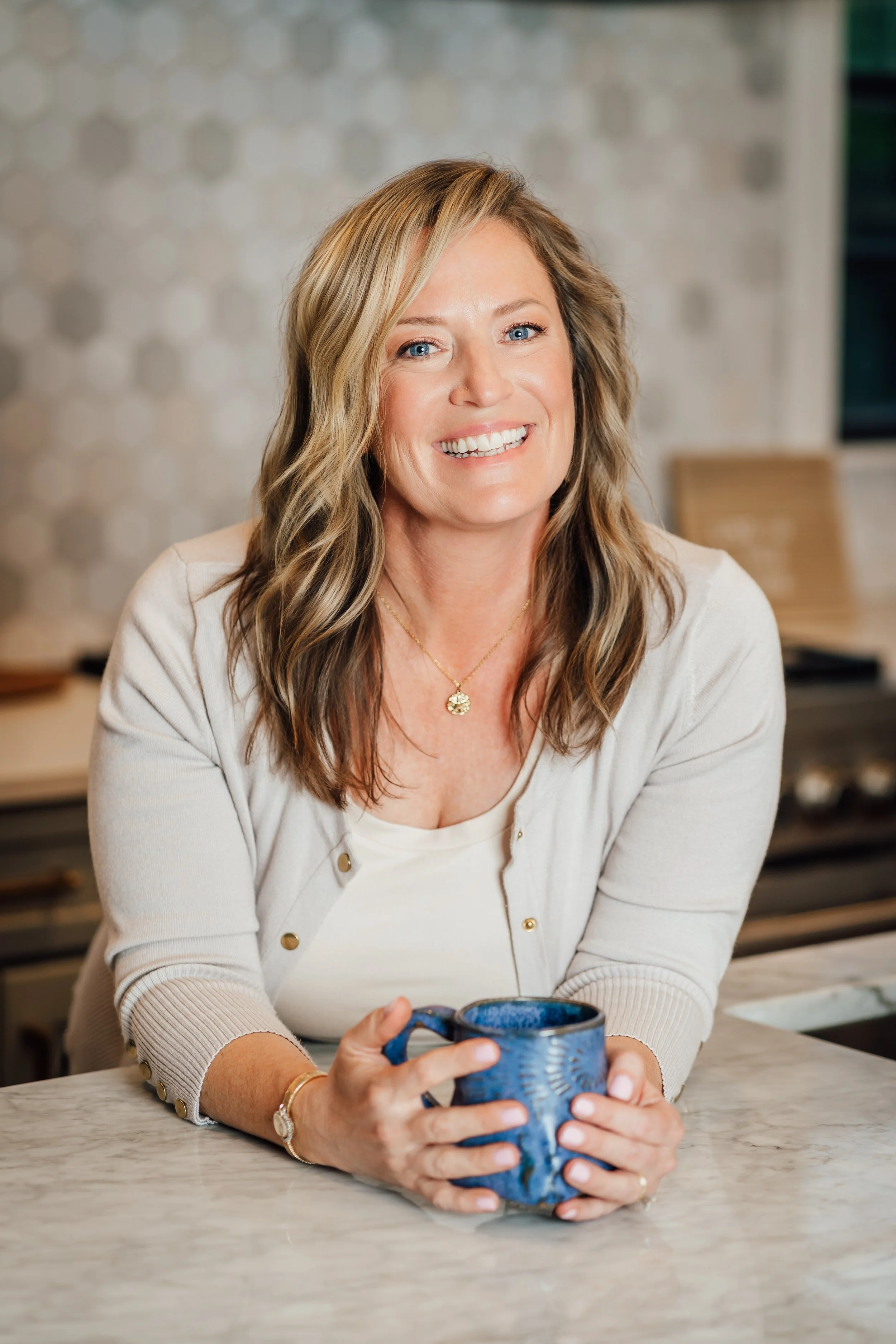 A woman with blonde wavy hair and blue eyes sitting at a marble table, holding a blue ceramic mug, smiling, in a modern kitchen with a textured wall background.