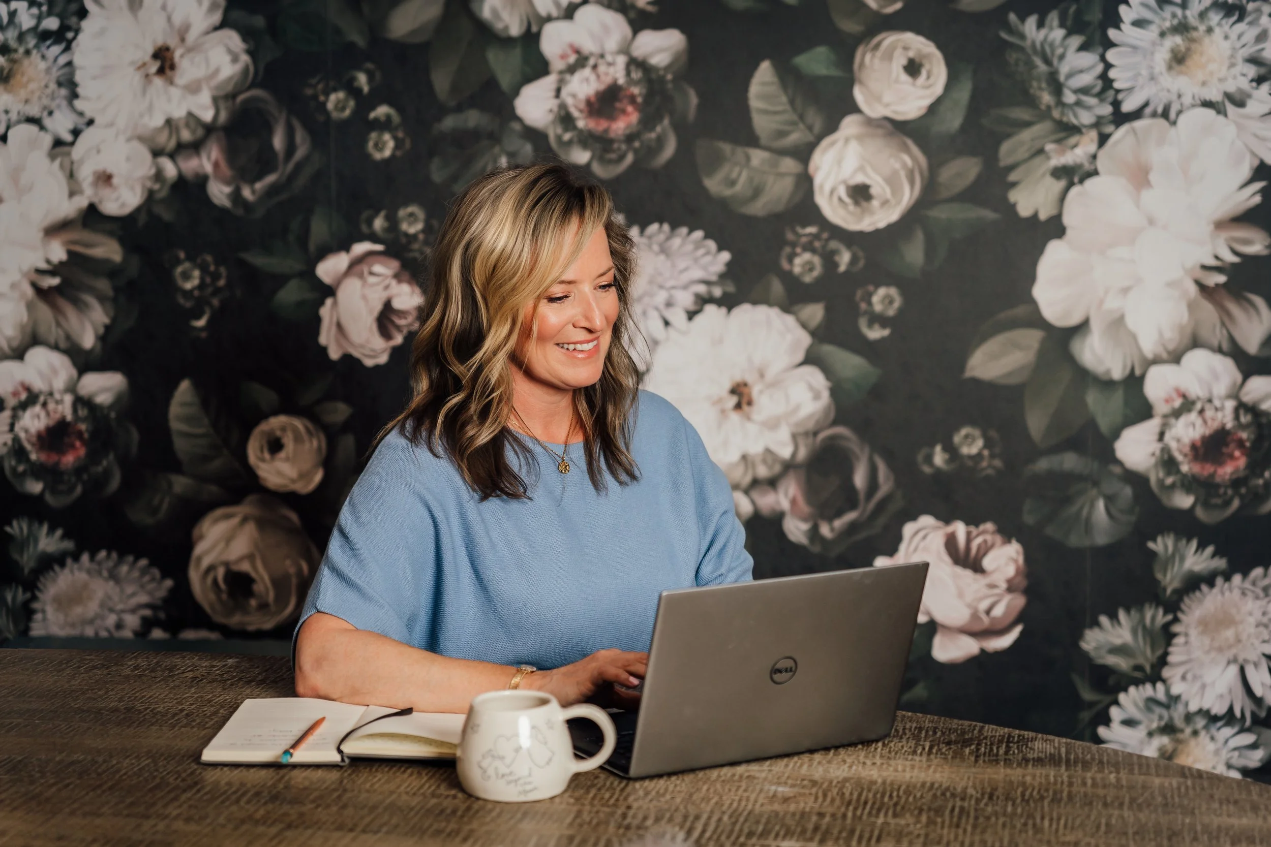 A woman with blonde and brown wavy hair, wearing a light blue shirt, sitting at a dark wooden table with a silver Dell laptop, a white mug, a notebook, and a pen, in front of a floral wallpaper background.
