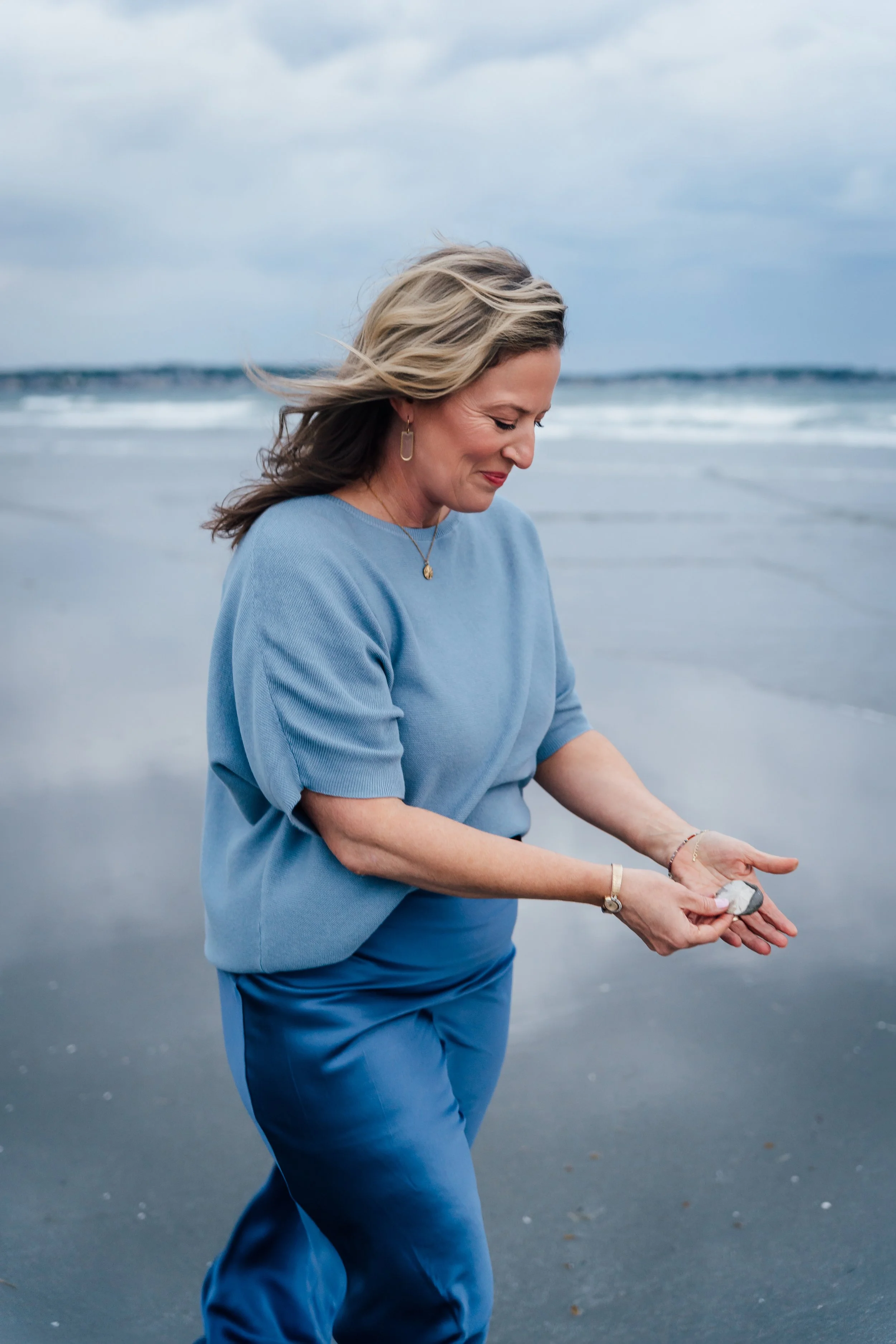 A woman with blonde hair and a blue sweater stands on a beach with wind-blown hair, holding a shell in her hands, with the ocean and cloudy sky in the background.