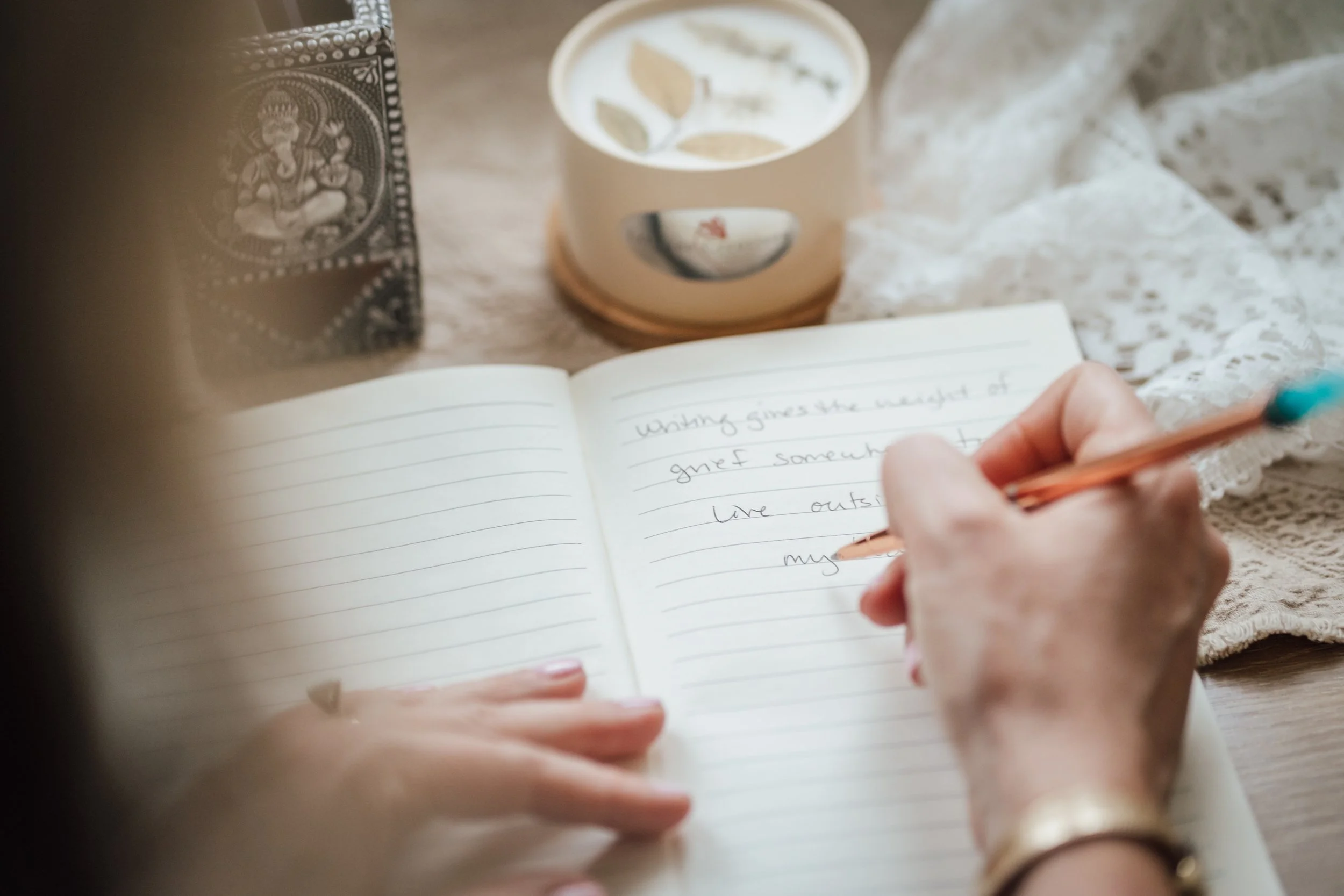 A person writes in a lined notebook with a orange pen, with a cup of tea or coffee, and a patterned black and white bandana on the table.