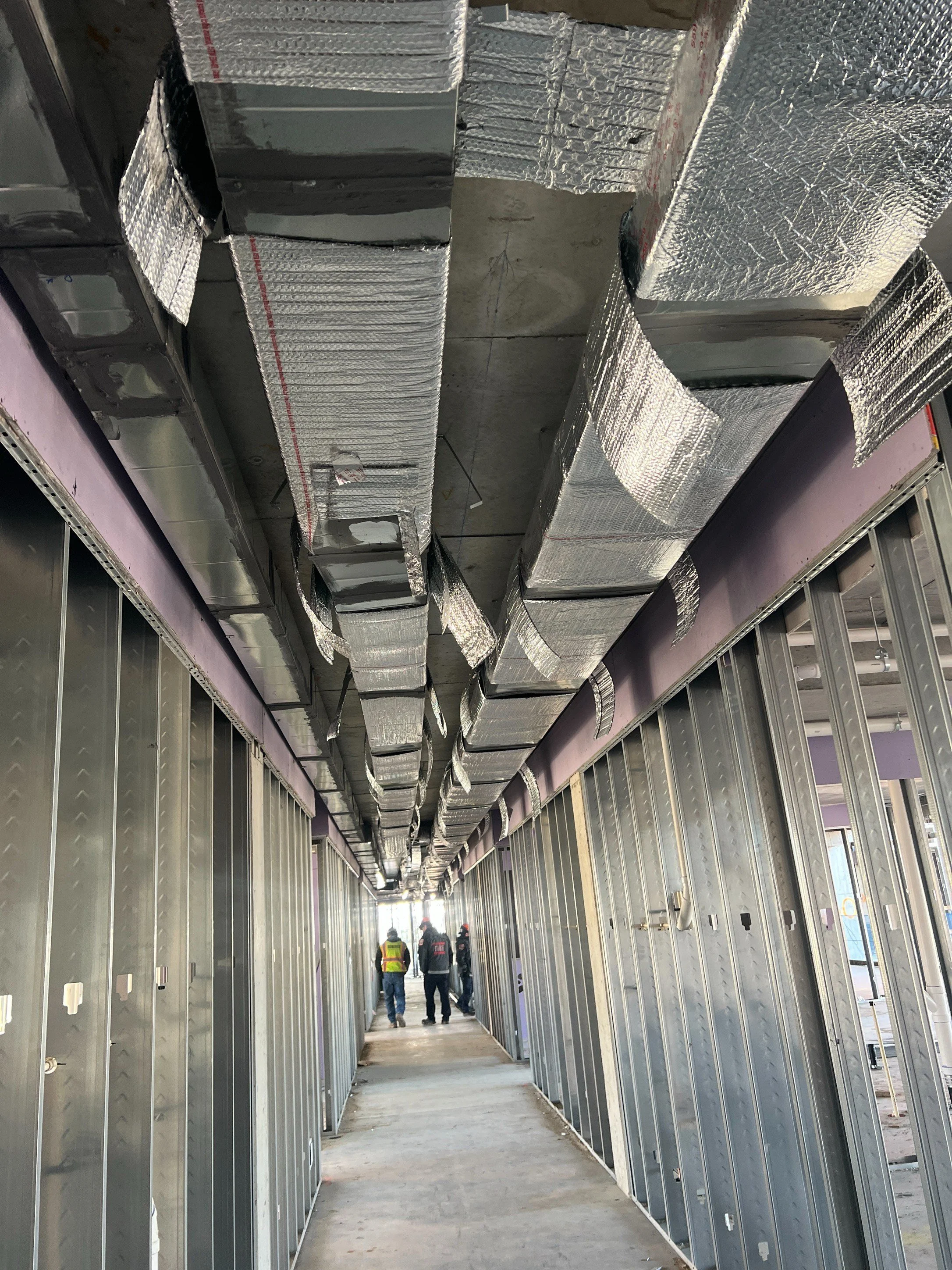 Construction workers walking through a hallway under construction with exposed metal framing and ductwork on the ceiling.