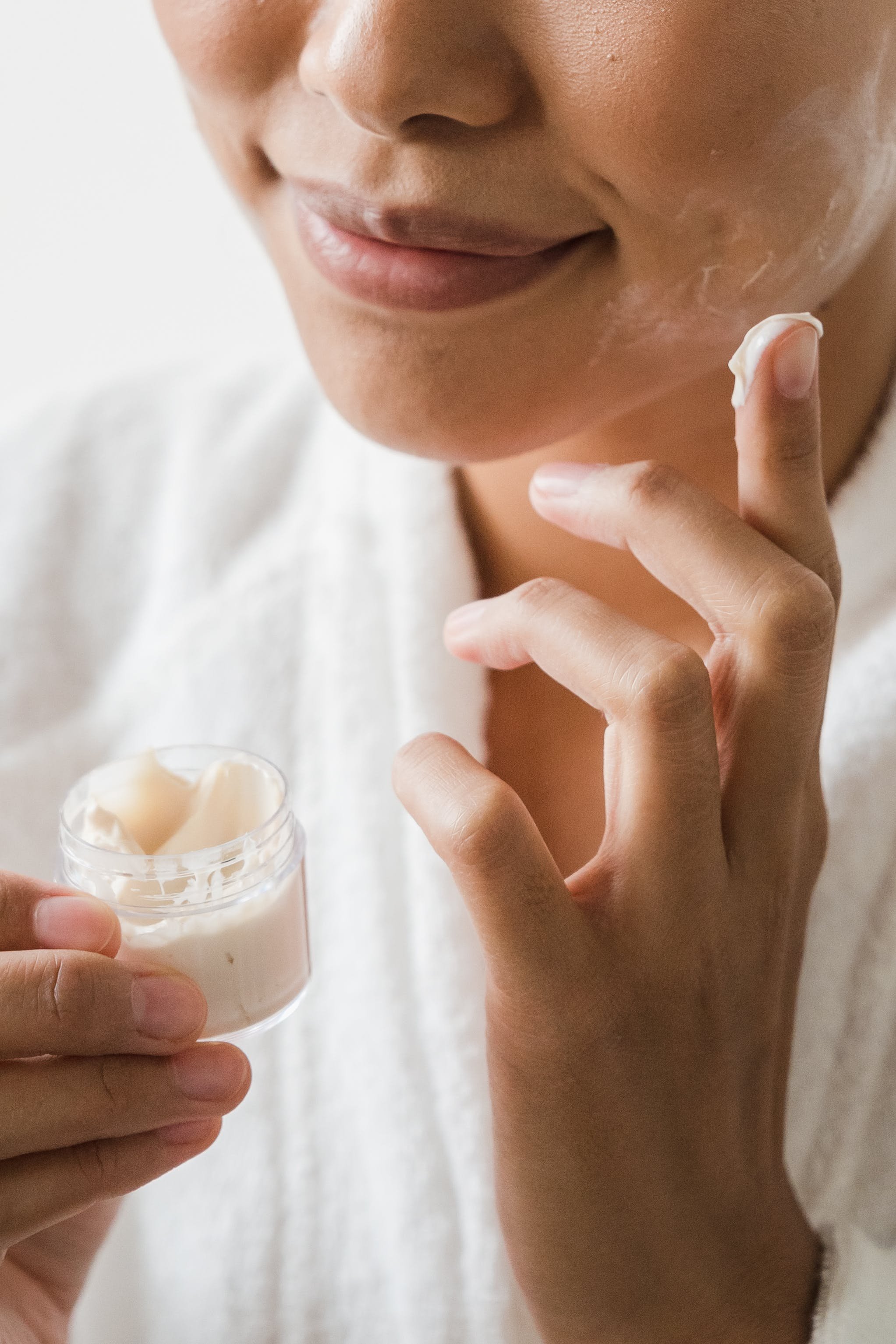 A woman applying moisturizer to her face, holding a small jar of cream in her other hand.