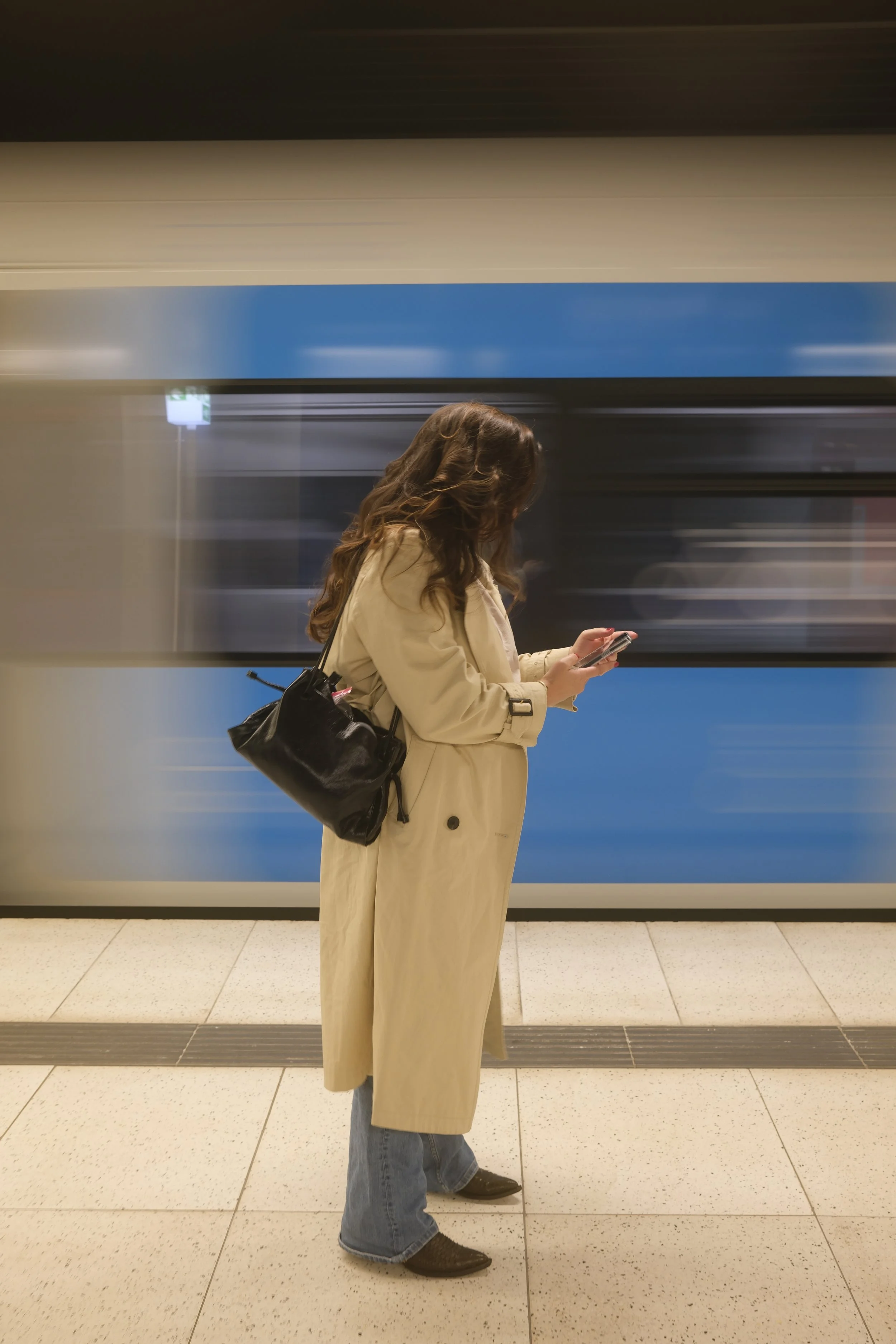Eine Frau mit braunen lockigen Haaren, die eine beige Trenchcoat und Jeans trägt, steht in einem U-Bahn-Station und schaut auf ihr Smartphone.