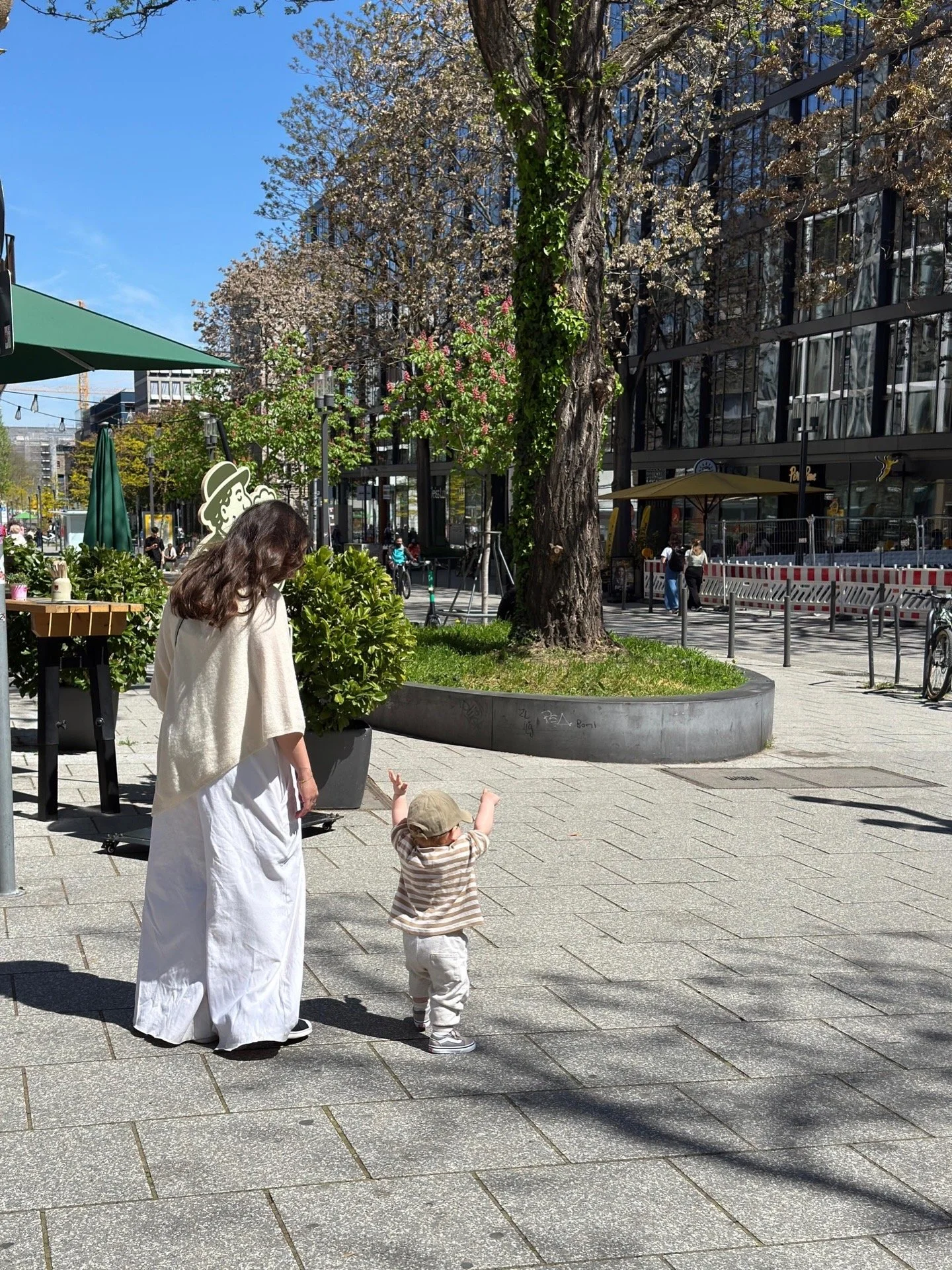 Eine Frau in einem weißen Kleid und eine kleine Person in gestreiftem Shirt und Kappe spielen auf einem Gehweg in der Stadt. Im Hintergrund sind Bäume, Büros und ein Straßensperrzaun zu sehen.