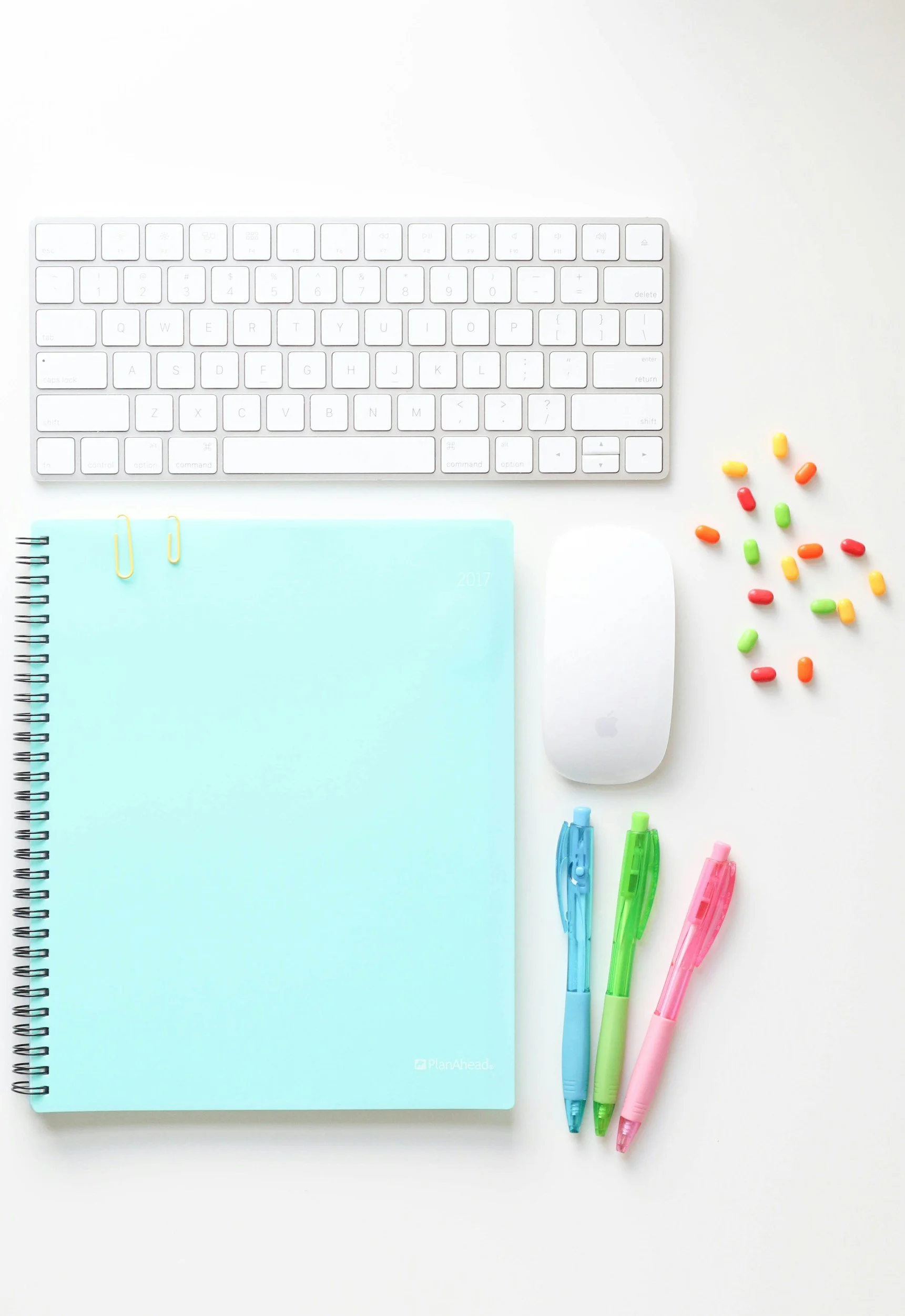 A white computer keyboard, a blue spiral notebook, three colorful pens, a white mouse, a scattered collection of colorful pills or candies, and two highlighters on a white surface.
