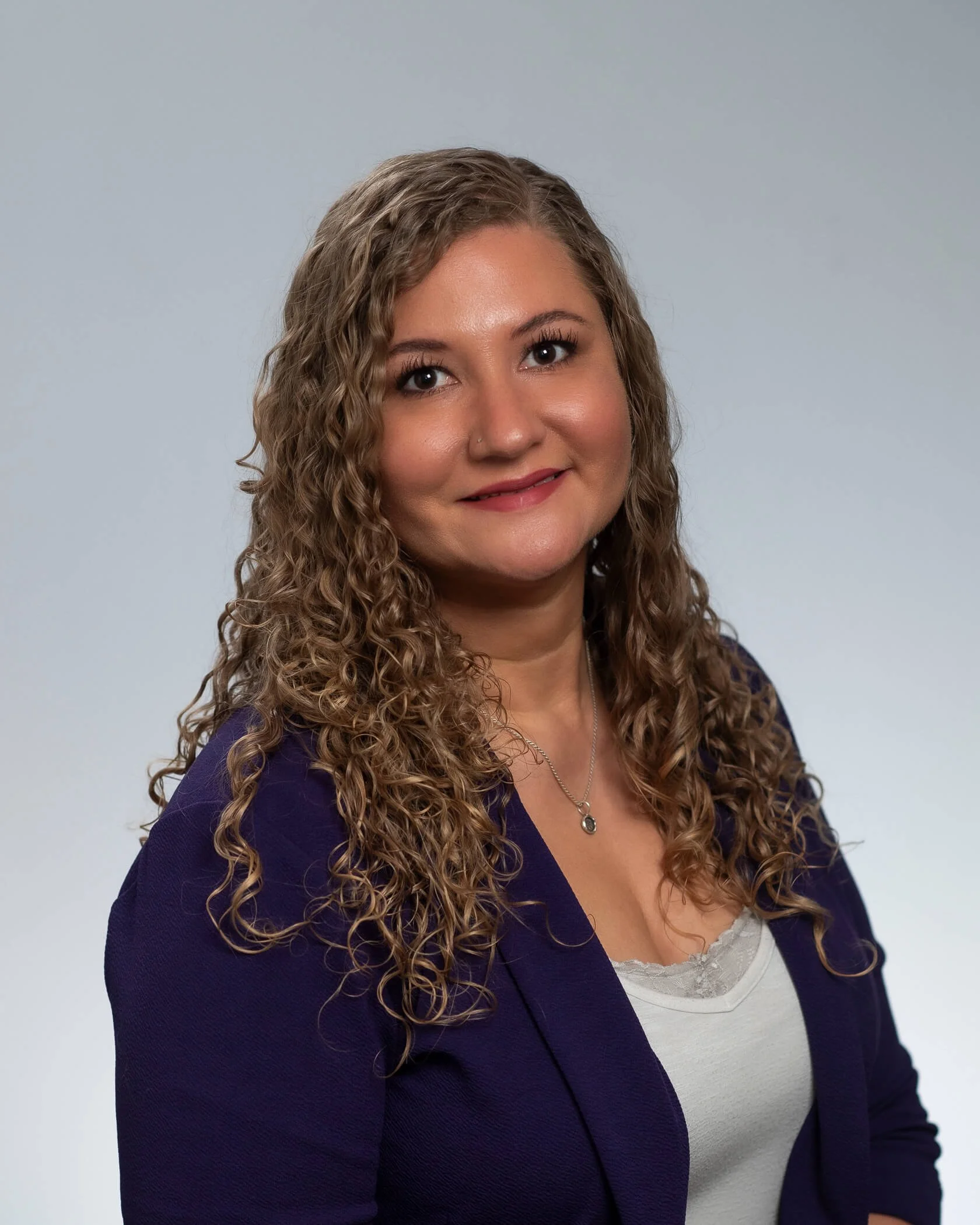 Close-up of a woman with curly hair wearing a purple blazer and a white lace-trimmed top, standing against a plain light gray background.