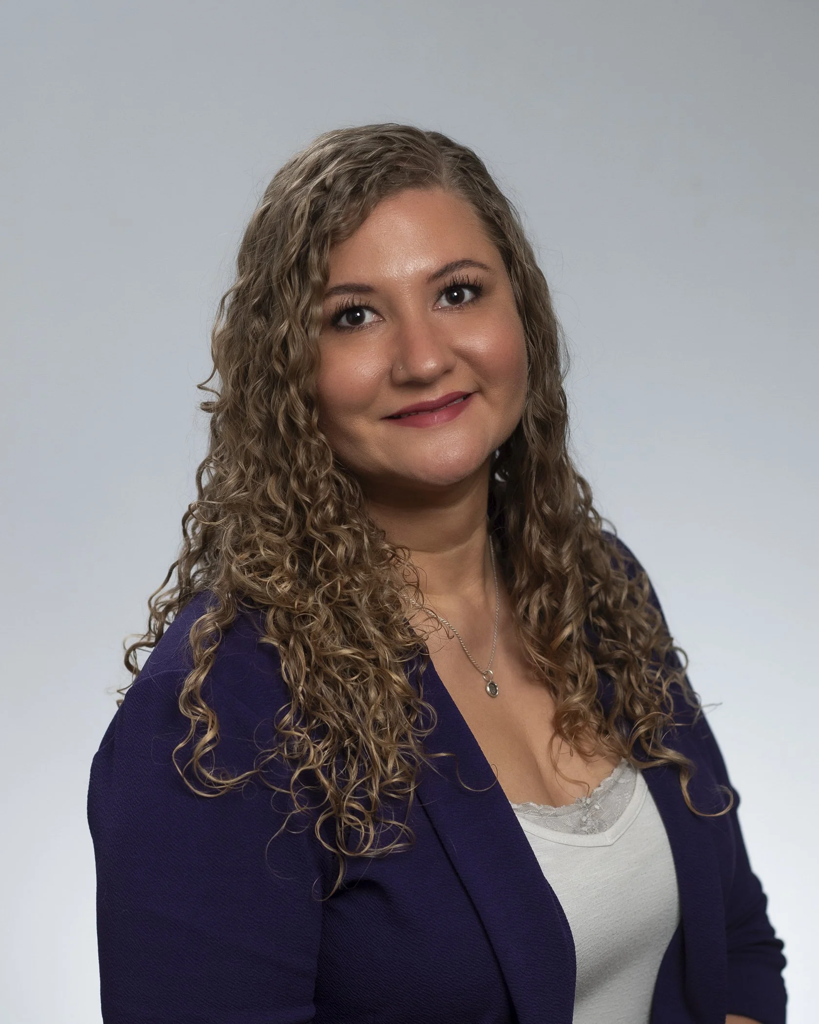 A woman with long, curly brown hair, wearing a purple blazer over a white top with lace, standing against a plain light gray background.