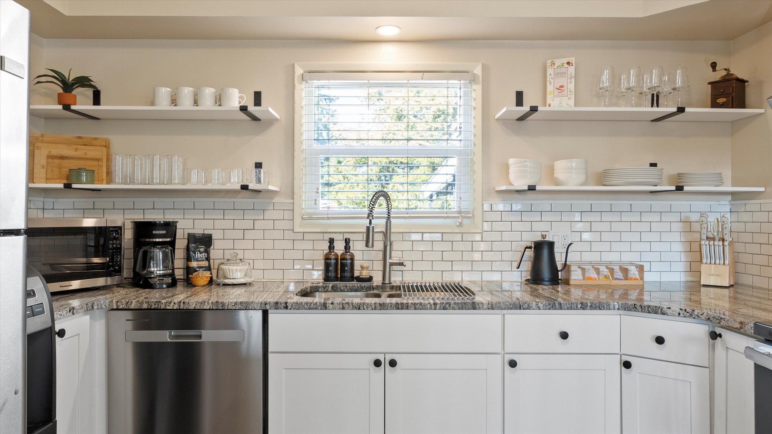 Kitchen with open shelves, white subway tile backsplash, window above sink, black kettle, coffee machine, mug, and glassware.