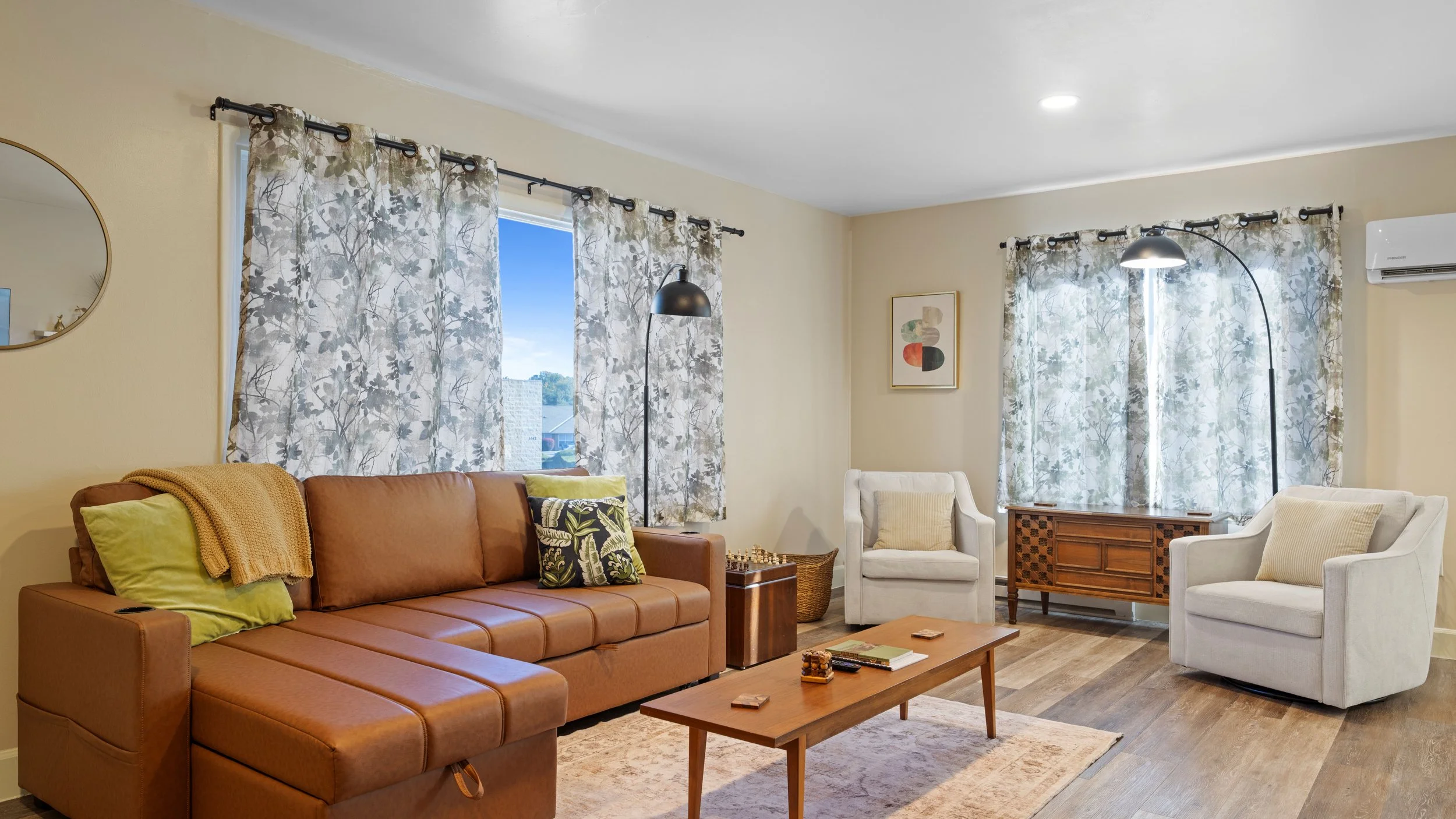 Living room with beige walls, hardwood flooring, a brown leather couch with throw pillows, white armchairs, a wooden coffee table, and windows with patterned curtains.