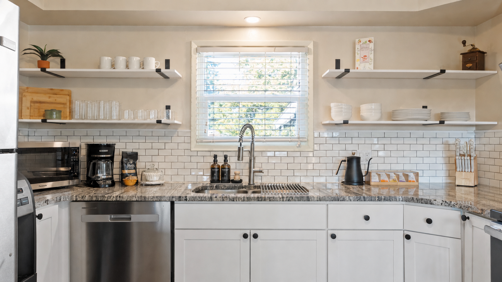 Modern kitchen with white cabinets, gray granite countertop, stainless steel dishwasher, open shelving with white dishes and cups, window with blinds, coffee maker, kettle, and tea packet holder.