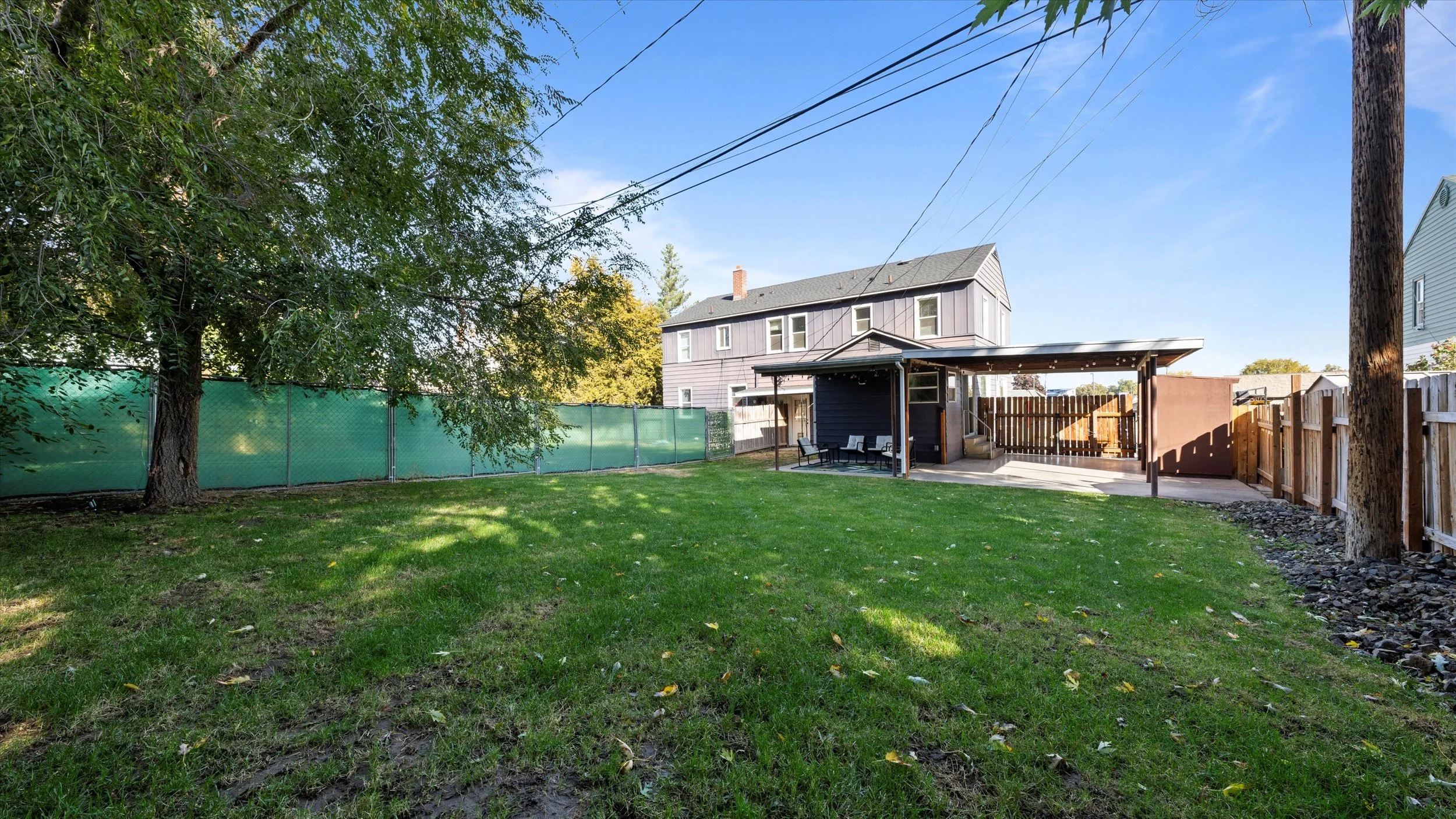 Backyard with green grass, a large tree, wooden fence, and a house with a covered patio and outdoor seating.