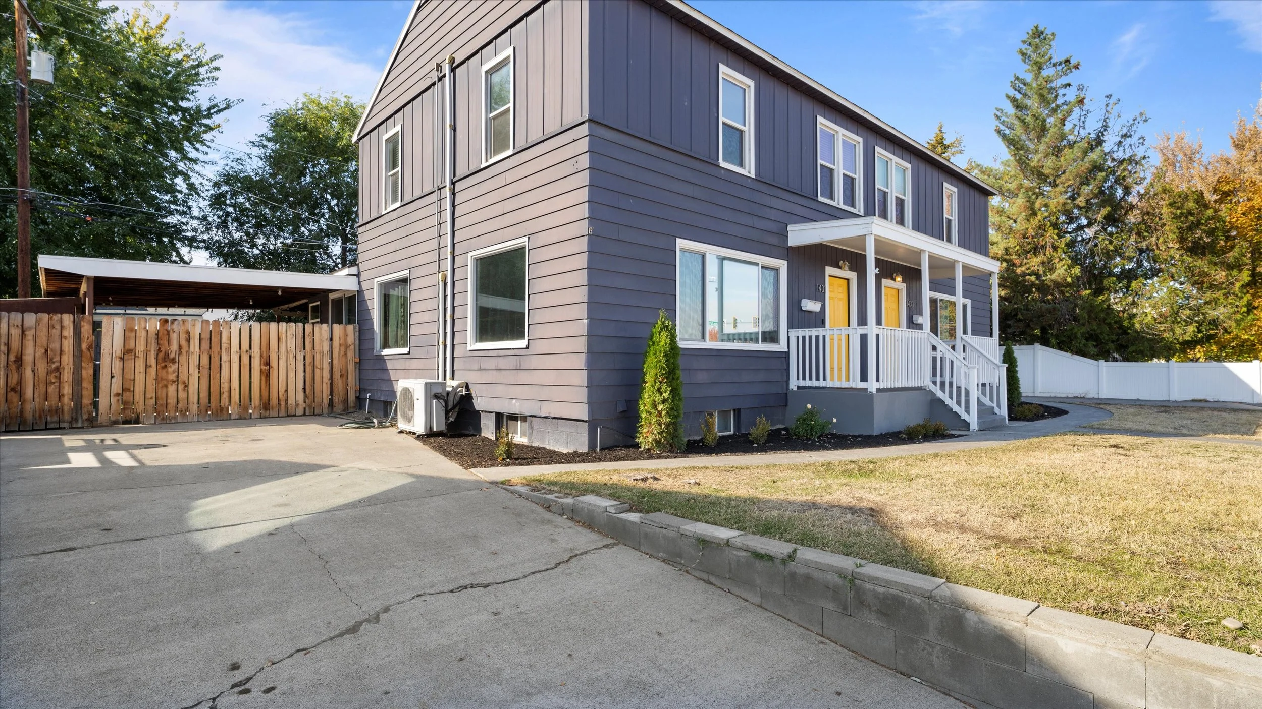 A two-story modern house with gray siding, yellow front doors, a small porch with white railing, and a concrete driveway, surrounded by a wooden fence and trees.