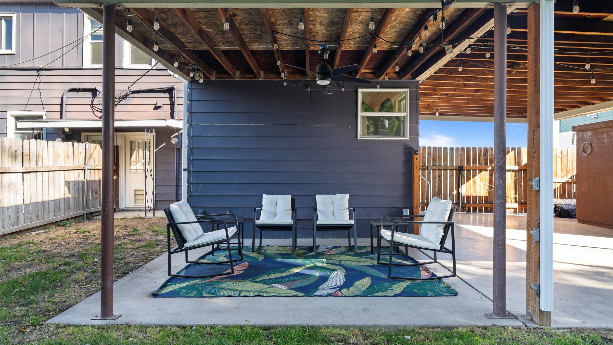 Image of a backyard patio with black metal chairs and cushions, a colorful outdoor rug, a wooden fence, and an overhanging roof with string lights.